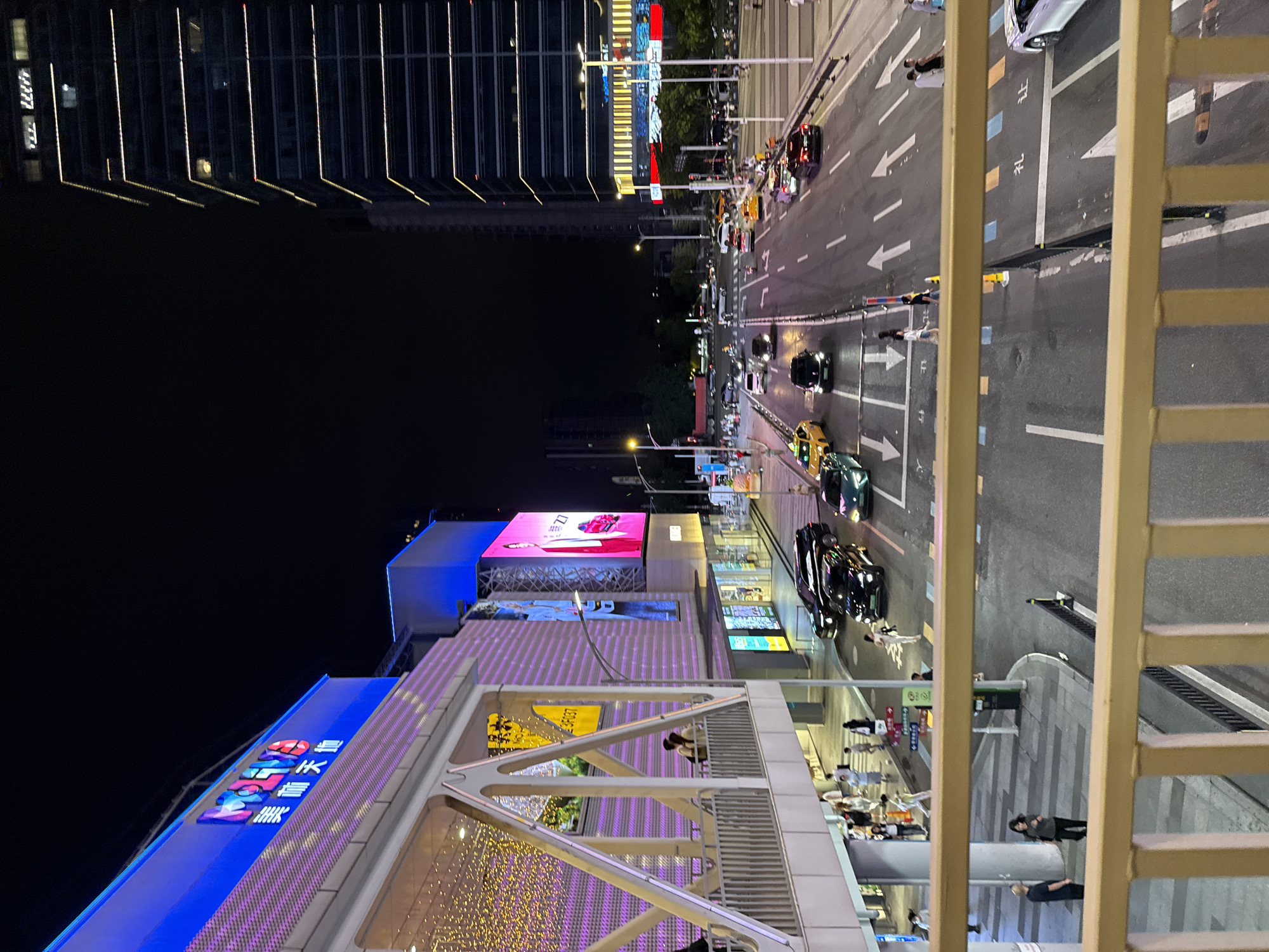 The image captures a nighttime urban scene in Tianhe District, China, from an elevated vantage point, possibly a pedestrian bridge. A multi-story commercial building, illuminated with blue and purple LED lights, dominates the left side of the frame. The building features a prominent sign with colorful, illuminated characters reading "MOLINO" and Chinese text below. A large digital billboard on the building displays an advertisement featuring a person and a red car.
Below the commercial building, a street bustles with traffic. Several cars, including black SUVs, a green sedan, and yellow taxis, are visible on the road. Pedestrians are seen walking on the sidewalks and crossing the street.
To the right, a tall, modern skyscraper with horizontal bands of yellow lighting stretches towards the dark night sky. The skyscraper's facade is segmented by these illuminated lines, creating a distinct geometric pattern. Streetlights and traffic signals are also present, contributing to the illuminated urban landscape. The overall atmosphere is one of a vibrant, commercial district at night.