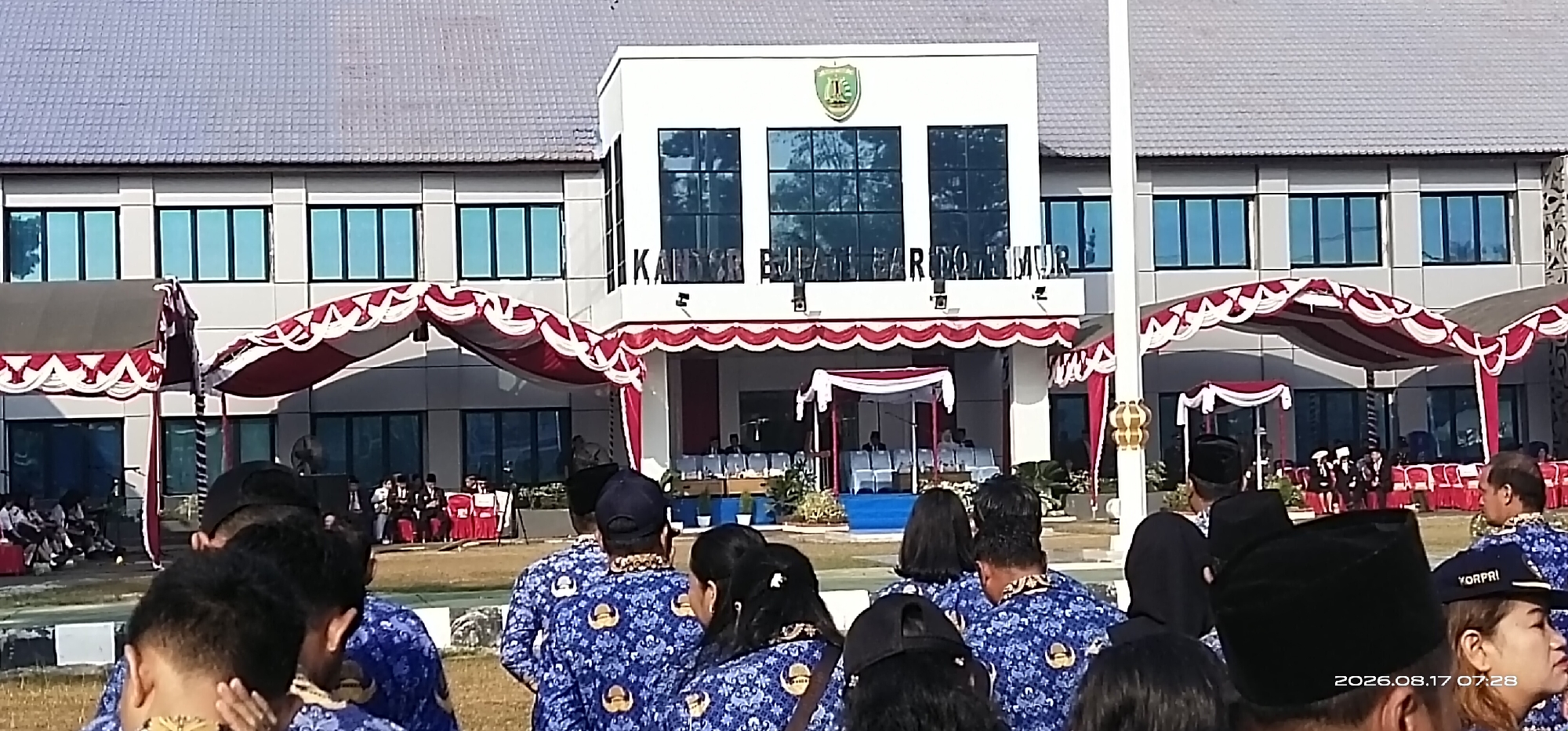 In Tamiang Layang, Indonesia, an outdoor ceremony is taking place in front of a modern, white building with a gray roof. The building features large, reflective windows and is adorned with a green emblem at the top center, displaying the text "KANTOR BJNPARTOLIMUR". Red and white striped awnings are set up in front of the building, and a white tent with a red trim is also visible. A crowd of people, many wearing blue patterned uniforms, are gathered on a grassy field. Some individuals are seated, while others are standing. A flagpole with decorative golden elements stands to the right of the frame. The lighting suggests it is daytime.