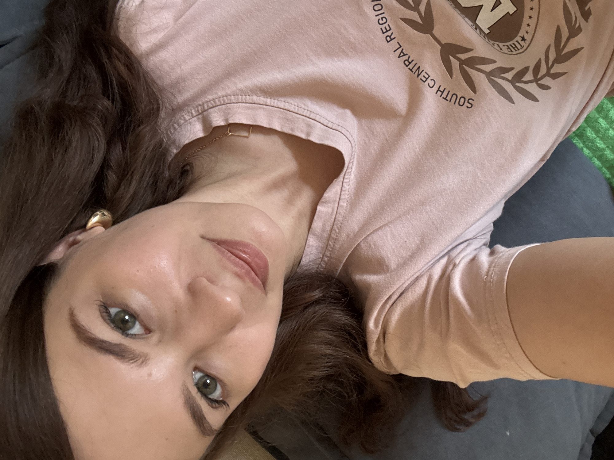 The media file is a close-up, overhead selfie of a young woman lying down. She has long, dark brown hair that falls around her face. Her skin is fair, and she has green eyes and well-defined eyebrows. She is wearing a light pink t-shirt with a graphic on the front. The graphic appears to be a circular emblem with text around it, including "SOUTH CENTRAL REGION" and "THE". She is also wearing a simple gold necklace with a square pendant and a large gold hoop earring in her left ear. The background is dark and appears to be a sofa or bed. The lighting suggests it might be daytime, possibly indoors. Her expression is relaxed and a little playful, with her head tilted and a slight smile. The overall mood is casual and intimate.
