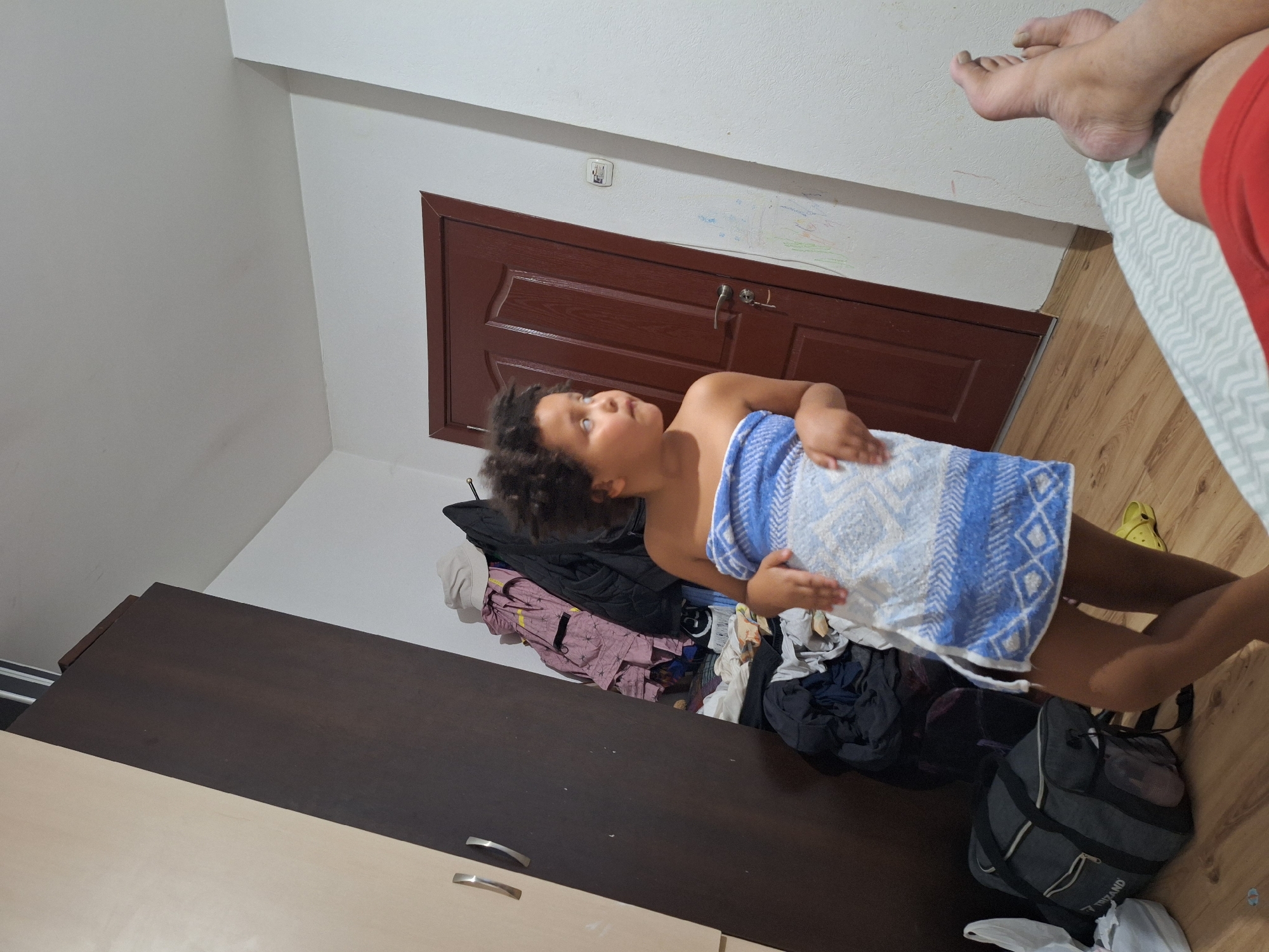 The image shows a young child wrapped in a blue and white patterned towel, looking upwards with curiosity. The child is positioned in what appears to be a bedroom. To the left of the child is a dark brown wardrobe with two handles. On top of the wardrobe, a pile of clothes is visible, including a pink garment with a geometric pattern, a black jacket, and other folded items. To the right of the child, a wooden floor is visible, along with a white and light blue patterned bedspread. A pair of feet, likely belonging to an adult, are resting on the bedspread, wearing red shorts. Further to the right, a yellow Croc shoe is also on the floor. In the background, a dark brown wooden door with a silver handle is visible. Above the door, a light switch and some colorful scribbles on the wall are present. The overall lighting suggests it is daytime. The scene appears to be in a residential setting, likely a home in Blagoevgrad, Bulgaria. There is no visible text in the image.