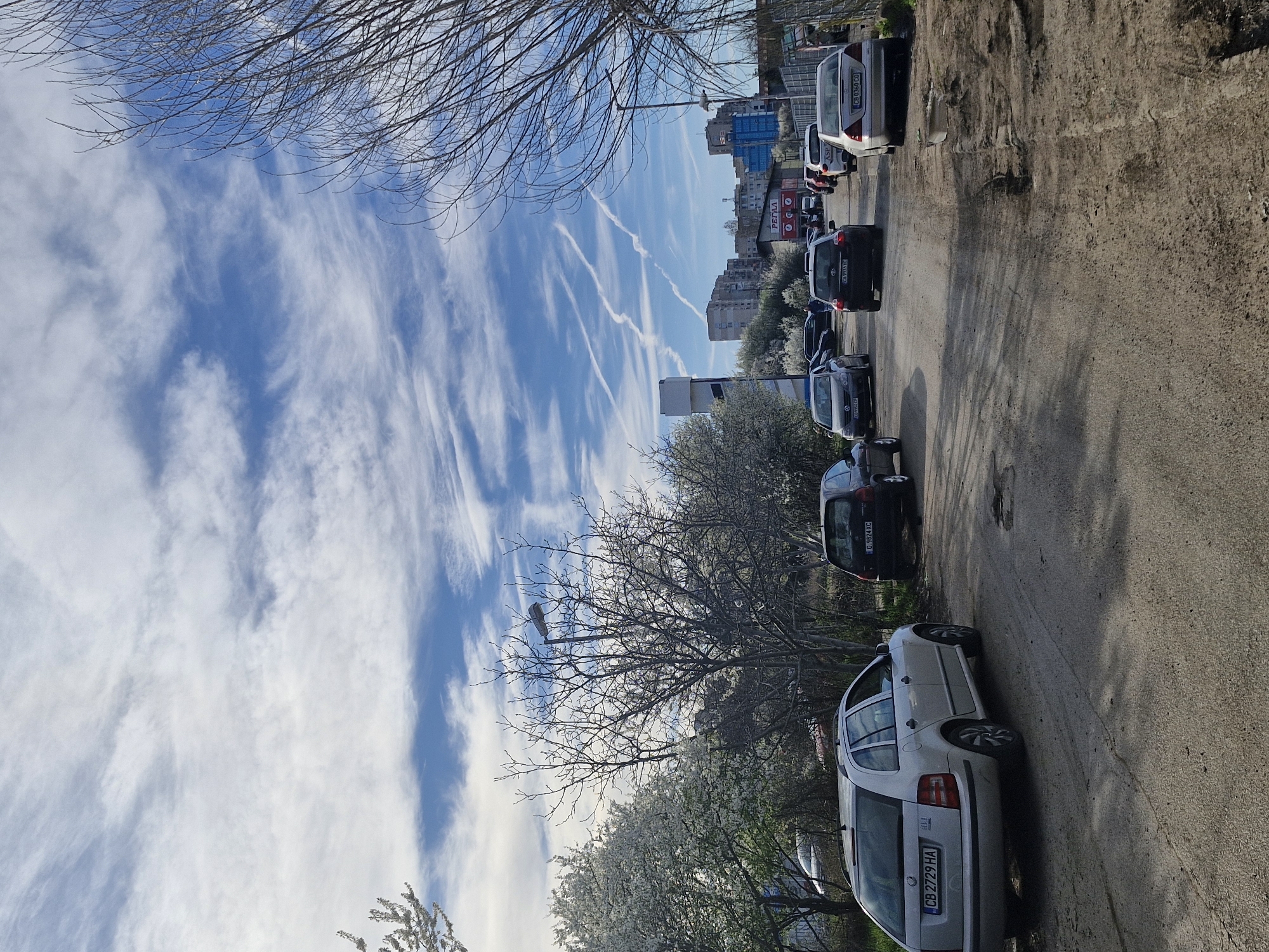The image depicts a street in Sofia, Bulgaria, lined with parked cars. The sky is blue with streaky clouds and contrails from aircraft. Bare trees and flowering bushes are visible along the side of the street. Buildings are visible in the distance.