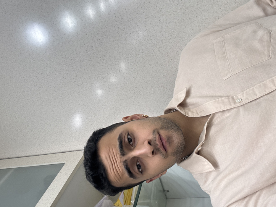 A selfie of a man with dark hair and eyes, a fair complexion, and a light beard. He is wearing a beige button-down shirt and a silver chain. The man's expression is slightly furrowed. The background appears to be a room with white walls and ceiling, with fluorescent lighting reflected on the surfaces. The camera angle is slightly tilted.