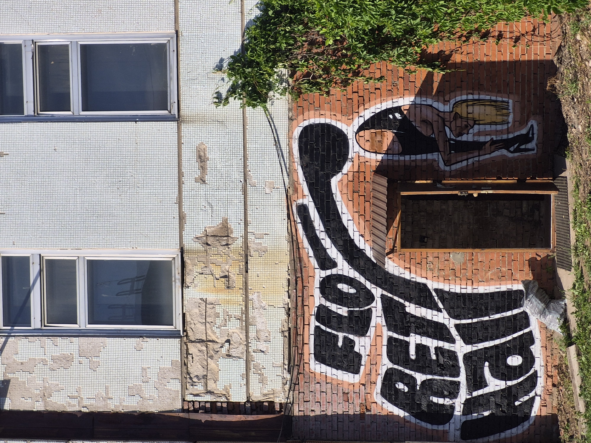 The image depicts the side of a building in what appears to be an urban setting, in the city of Unknown, Unknown. The building has two distinct sections: one is covered in pale blue tiles with two windows, and the other is a brick wall with graffiti and an open door or window space near the top. On the brick wall, there is graffiti featuring a figure and large black and white lettering with the word "GENOP". Green foliage grows on top of the brick wall section and around the right side. The blue-tiled section shows signs of deterioration. The photograph is taken from an overhead perspective.