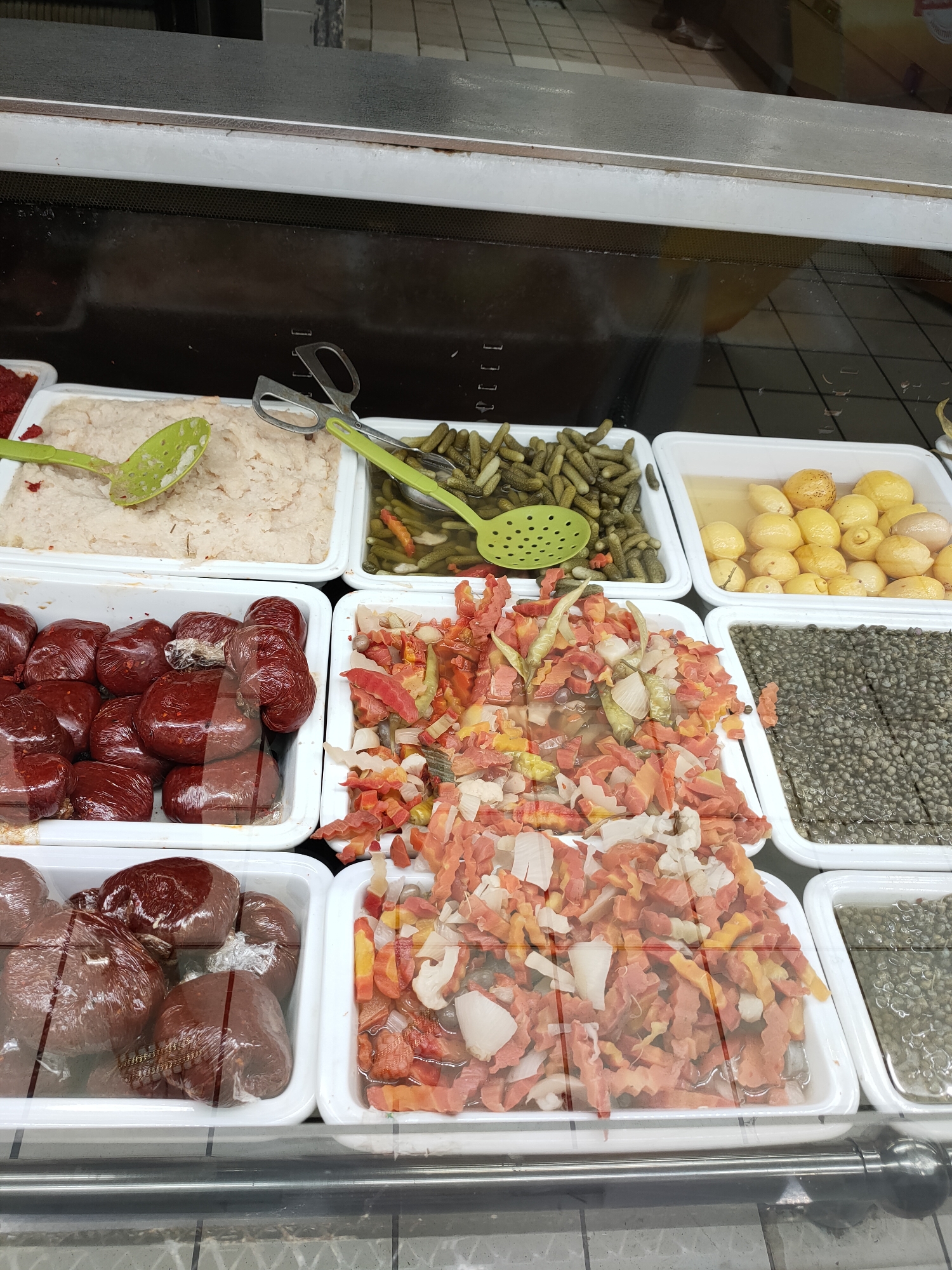 In a brightly lit refrigerated display case, a variety of preserved foods are presented in white rectangular trays. The items include a creamy, pale spread in the top left, likely a dip or pâté, served with a green slotted spoon. Next to it are small, pickled gherkins in brine, also with a green slotted spoon and a pair of metal tongs. To the right of the gherkins are small, whole lemons, preserved in a light yellow liquid.
Below these, in the front row, are several trays of chopped and mixed ingredients. The leftmost tray contains what appear to be small, dark red, individually wrapped food items, possibly sausages or meat preparations. Adjacent to this is a tray filled with a colorful mix of chopped vegetables, predominantly orange and white, with hints of red and green, possibly a salad or relish. To the right of the vegetable mix, another tray holds small, dark green, round objects, likely capers, in brine. Finally, in the bottom right, a tray contains more of these capers, with some other preserved items visible. The overall scene suggests a deli counter or a section of a grocery store specializing in preserved goods. There are no people visible in the frame. The lighting appears to be artificial, typical of an indoor commercial setting.