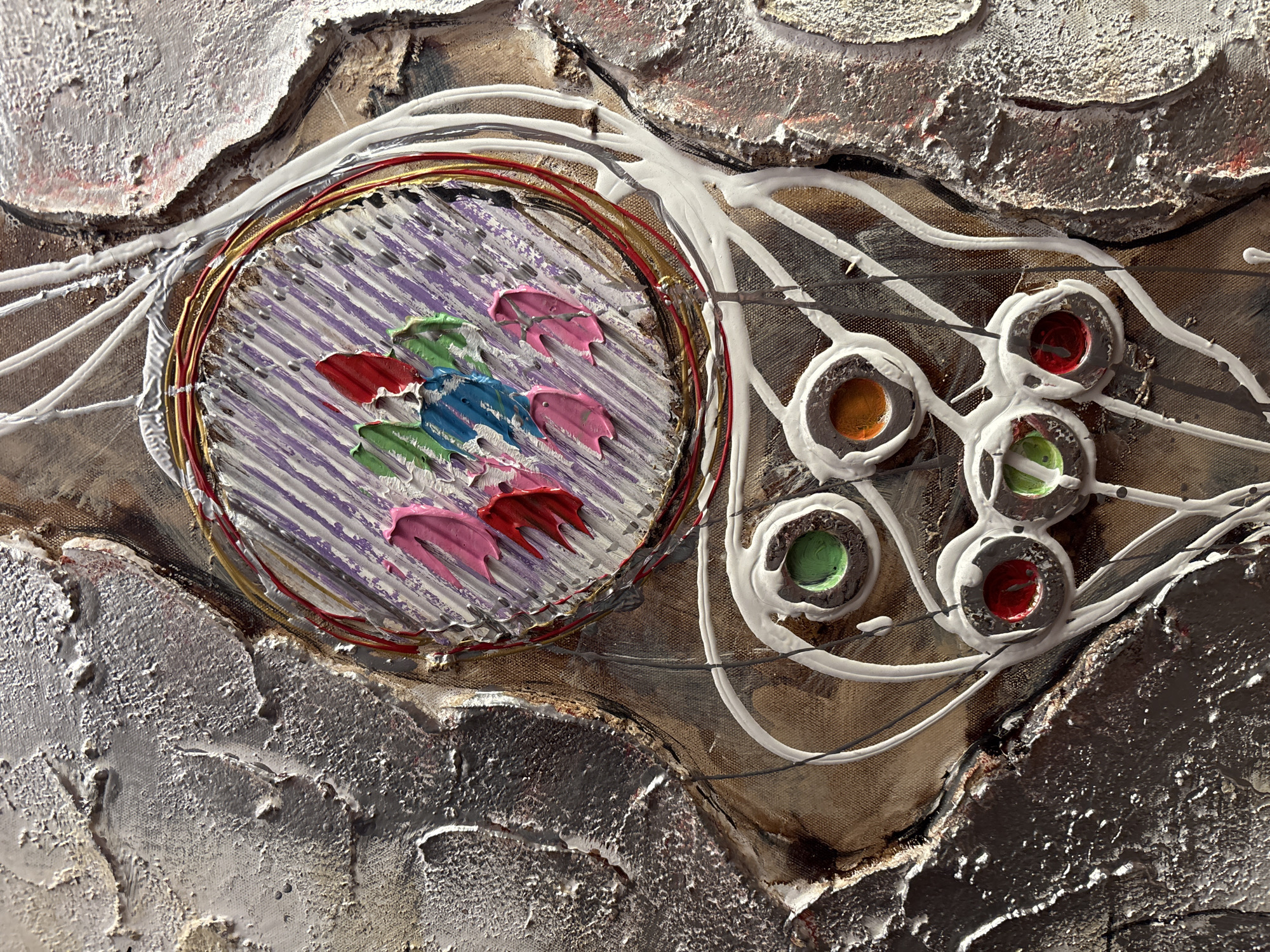 This is a close-up, abstract photograph of a textured artwork. The artwork features a variety of materials and techniques, including impasto paint, wire, and possibly some sort of resin or molded material.
The central subject is a circular element with concentric rings of wire, some appearing metallic gold and red. Inside this circle, there are parallel lines of purple paint, overlaid with thick, raised strokes of pink, blue, and green paint, giving the impression of small, abstract shapes or figures.
To the right of the central circle, there are several smaller, circular indentations filled with different colored substances. These are outlined with thick white paint, creating a drip-like effect. The colors visible in these circles are orange, red, and green.
The background of the artwork is highly textured, with areas of thick, white, stucco-like material that appears almost sculptural, interspersed with rougher, darker, and brown textured surfaces. Thin, dark lines are visible beneath some of the white paint, suggesting underlying drawings or wire elements.
The lighting suggests the artwork is illuminated from the side, casting shadows and highlighting the three-dimensional textures. There is no discernible text in the image. The overall impression is one of organic forms and vibrant, tactile surfaces.