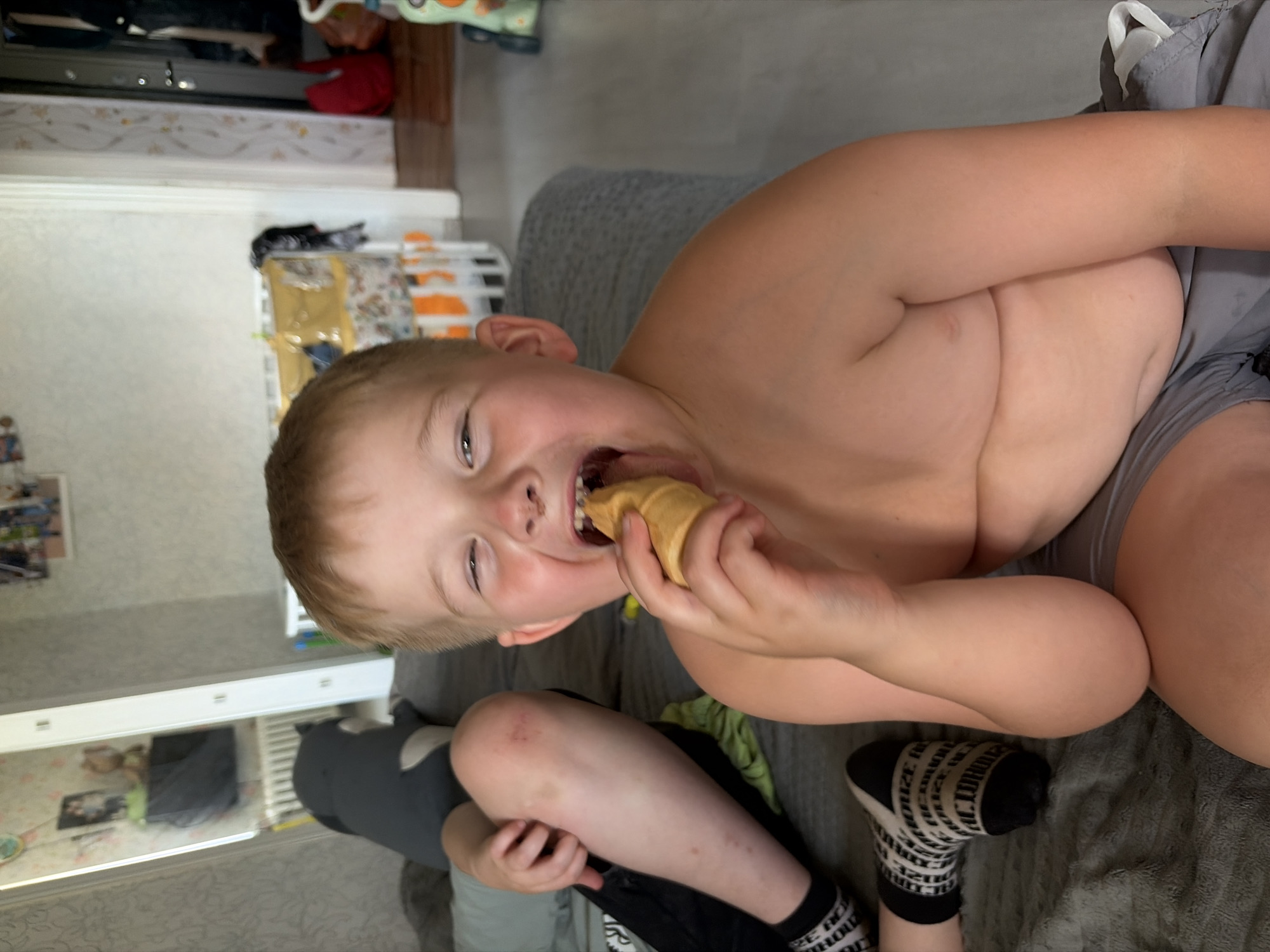 A boy is seen in a close-up shot, with his torso and head filling most of the frame. He is shirtless and wearing gray shorts. The boy is holding an ice cream cone up to his mouth and appears to be taking a bite, with his mouth open and eyes looking towards the camera. His expression suggests he is enjoying the treat. He has light brown hair and fair skin.
In the background, part of a couch with a gray textured fabric is visible. The scene appears to be indoors, possibly in a living room or bedroom, given the presence of furniture and what looks like a shelf with various items. The lighting suggests it might be daytime, but there are no clear indicators of the time of day.
Below the boy's torso, another person's legs are visible, wearing black shorts and black and white patterned socks. One of the socks has visible text in black lettering against a white background, which reads "LONDON." The legs are bent, with the person sitting or reclining.
The overall mood of the image is casual and domestic, capturing a candid moment of a child enjoying a snack. The setting appears to be in Russia, given the location context.