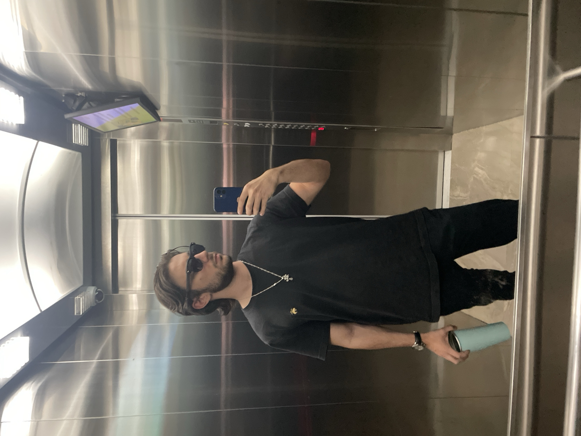 A man is taking a selfie in an elevator with metallic walls. He is wearing a black t-shirt, dark pants, and sunglasses. He has a beard and a silver chain with a pendant. In his left hand, he holds a blue cylindrical container. A display screen and a surveillance camera are visible in the elevator's upper portion. The floor is tiled. The location is Huixquilucan de Degollado, Mexico.