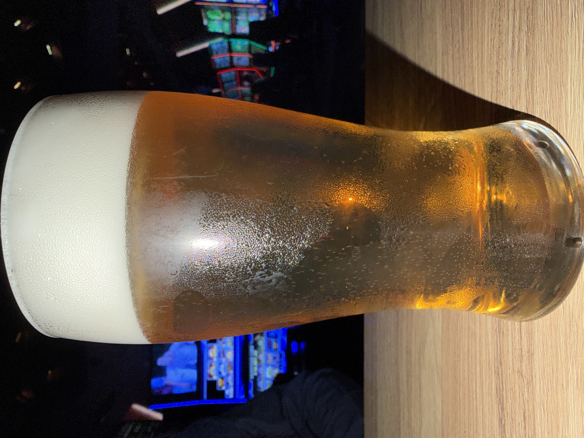 A close-up, eye-level shot shows a frosty glass of golden beer filled to the brim with a frothy white head. The glass is covered in condensation, and small bubbles rise from the bottom. The beer sits on a light brown wooden table. In the blurred background, colorful lights from what appears to be a casino or arcade are visible, with some shadowy figures in the distance. The scene is dimly lit, emphasizing the glow of the beer and the distant lights. There are no people clearly visible in the frame, and no specific landmarks of Burgas, Bulgaria, are discernible.