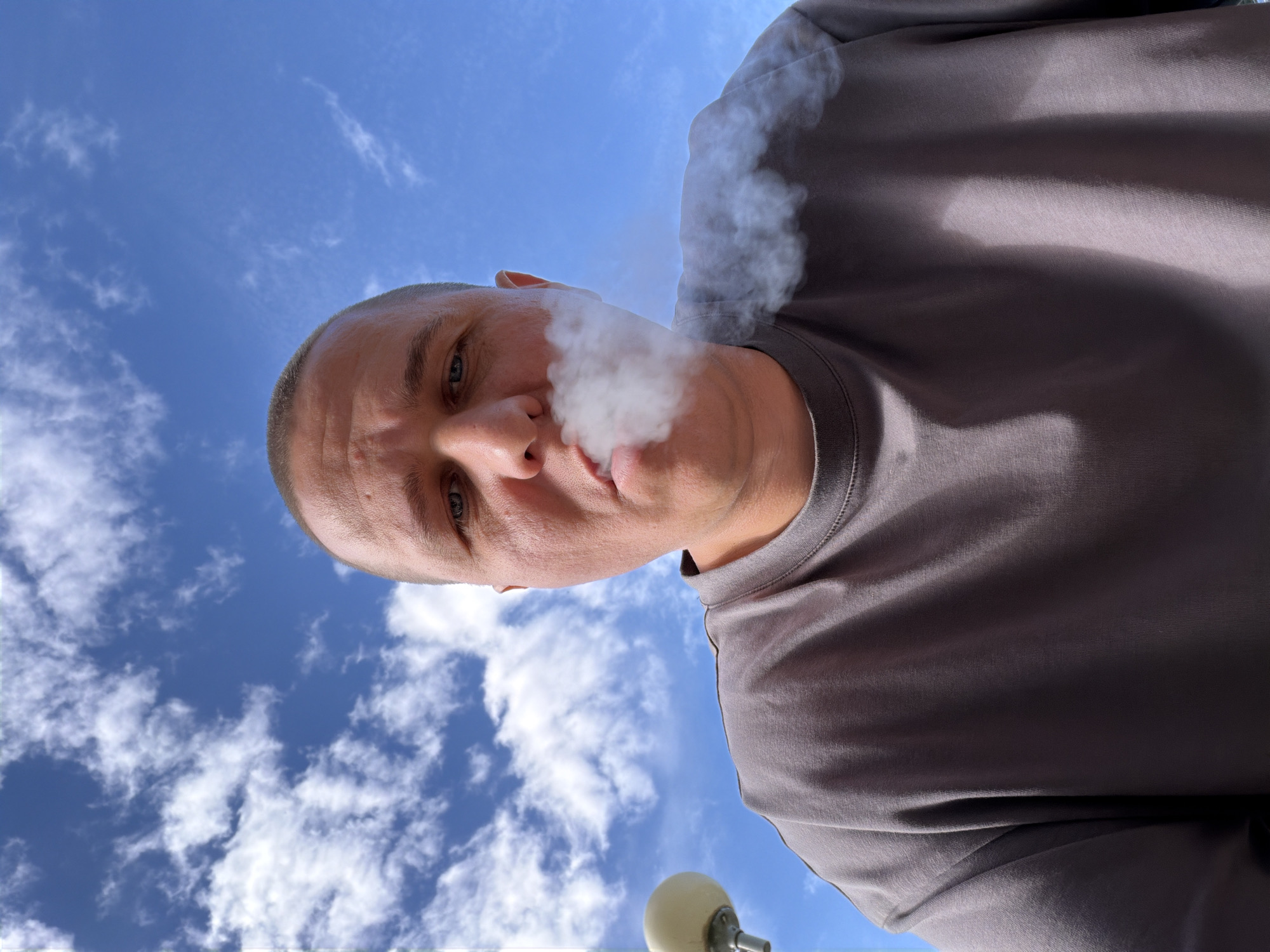The image is a close-up, low-angle selfie of a bald man exhaling a cloud of white vapor against a bright blue sky with scattered white clouds. The man is wearing a dark grey t-shirt. His face fills the lower half of the frame, with his eyes looking directly at the camera. To the left of the man's head, a portion of a white spherical object on a metallic pole is visible, which could be a street lamp. The scene appears to be outdoors on a clear day. The specific location in Minsk, Belarus is not discernible from the image.