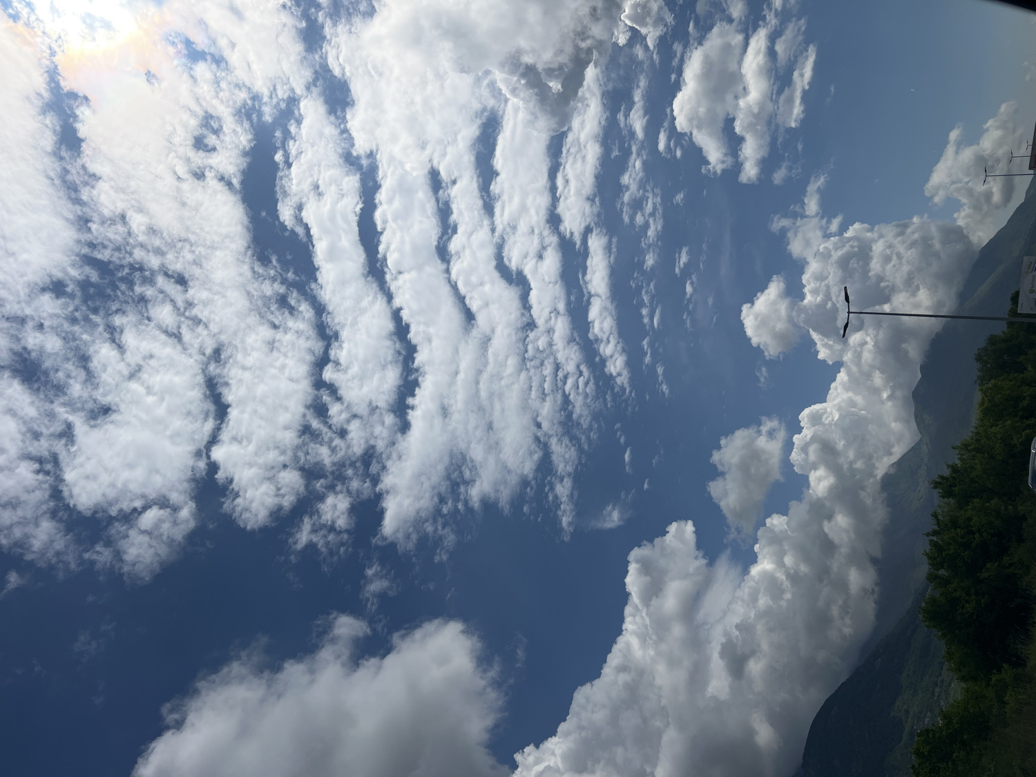 The image captures a vibrant sky filled with diverse cloud formations above a mountainous landscape, set in Kavač, Montenegro. The scene is dominated by a clear blue sky, partially obscured by clouds.
In the upper and left portions of the sky, delicate, elongated clouds stretch in parallel, rippled bands, reminiscent of altocumulus undulatus or cirrocumulus, with hints of a sun halo or glint in the upper left corner. These clouds appear lighter and more dispersed. Contrasting with these, large, fluffy cumulus clouds with significant vertical development are concentrated towards the right and bottom right of the frame, casting soft shadows upon themselves.
Below the expansive sky, the dark, forested slopes of mountains or large hills rise on the right side, partially enveloped or overshadowed by the larger cloud masses. Green tree foliage is visible at the very bottom right, at the base of these mountains.
A tall, dark streetlamp with two lights stands prominently on the right side, extending upwards into the sky. In the far background on the right, near the mountains, some indistinct man-made structures or signs are faintly visible, though no specific text is discernible.
The time of day appears to be daytime, likely mid-morning or afternoon, suggested by the bright natural light and the clear contrast between light and shadow. The weather is bright but very cloudy, with a mix of high-level and mid-level clouds. The overall mood is one of natural beauty and expansive calm.