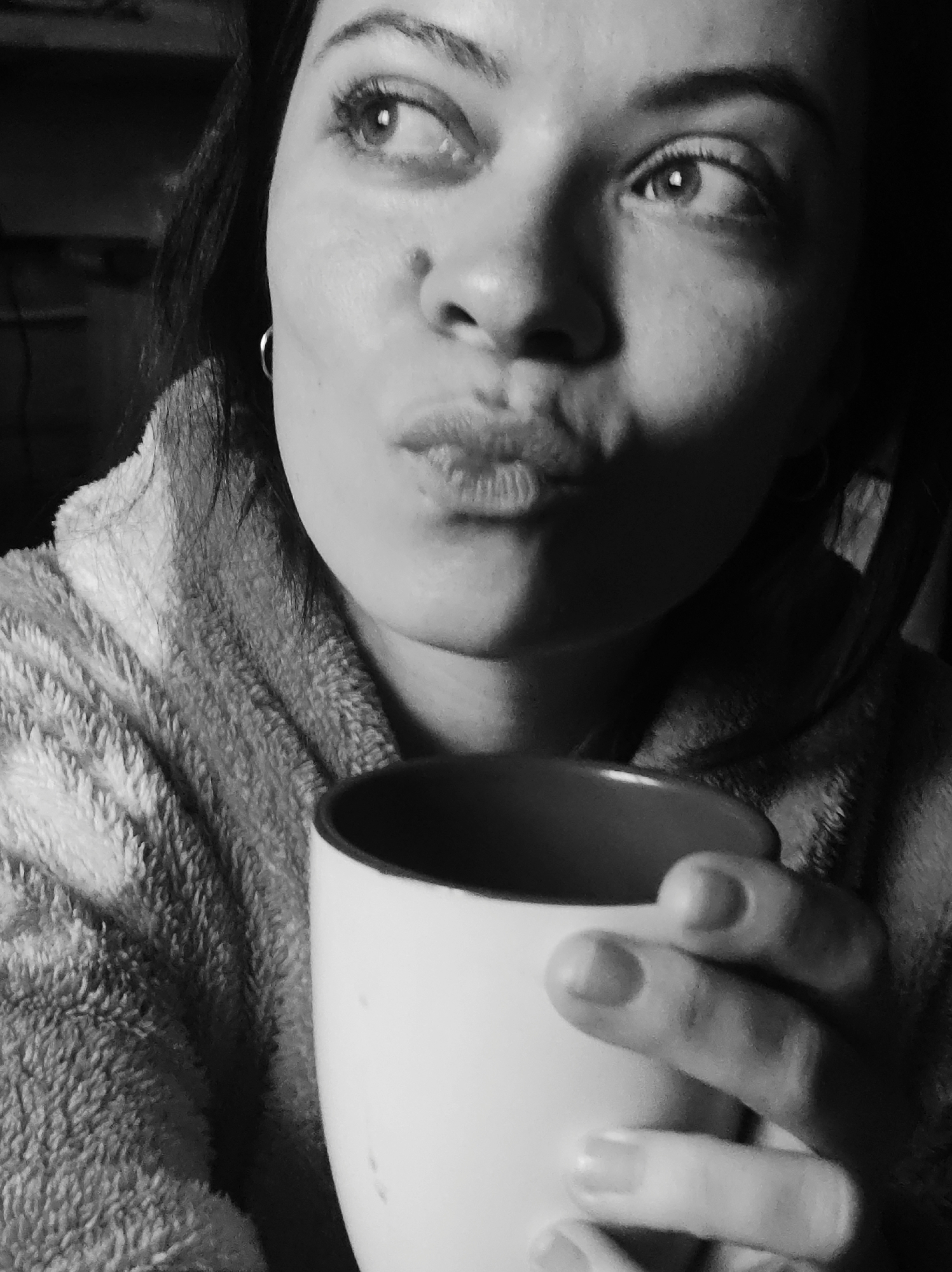 A grayscale medium shot depicts a woman holding a white mug with a dark interior. She is wearing a soft, textured robe. Her lips are pursed in a pouting expression, and her gaze is directed towards the upper left of the frame. She wears small hoop earrings. The location is unknown.