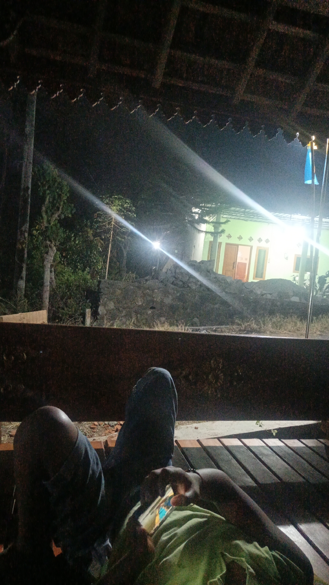 A person is lying on a wooden bench at night, holding a smartphone. They are wearing blue jeans and a light green shirt. In the background, a house with green walls and orange doors is visible, illuminated by bright lights. Two flags, one blue and one yellow, are planted in front of the house. The scene appears to be set in a rural area, possibly in Sambi, Indonesia. The environment is dark, with trees and foliage visible to the left of the house. The overall impression is one of a relaxed evening, with the person using their phone while enjoying the night air.