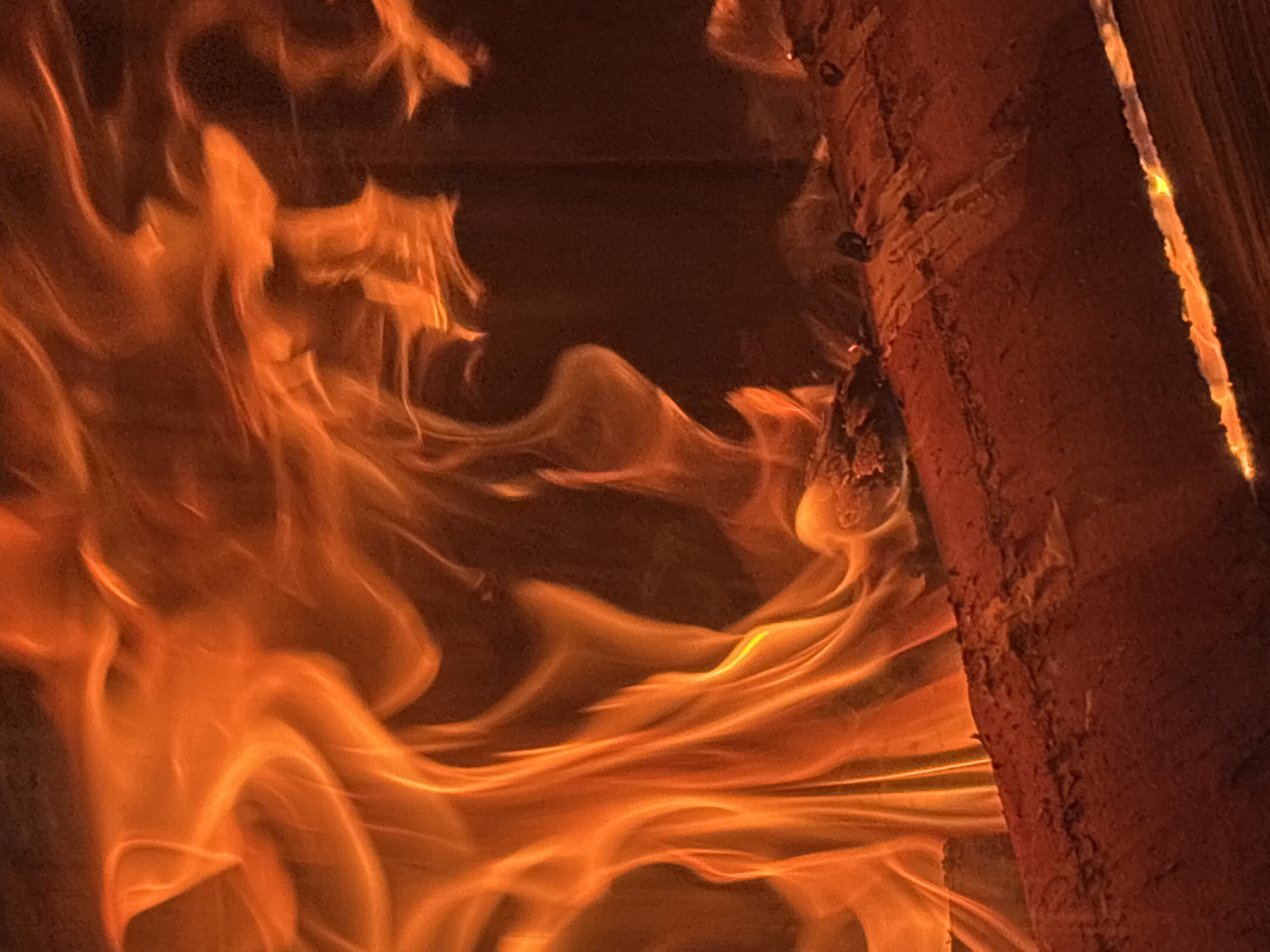 A close-up, low-angle shot of a fire burning. The flames are predominantly orange and yellow, with some wisps of white at their hottest points. The fire is consuming logs, parts of which are visible in the foreground and lower right of the image. The logs appear dark red and textured, with glowing embers scattered on their surface. The background is a dark, out-of-focus blur, suggesting the confines of a fireplace or hearth. The overall impression is one of intense heat and dynamic movement within the flames. There is no identifiable location or context suggesting Zvenigorod, Russia.
