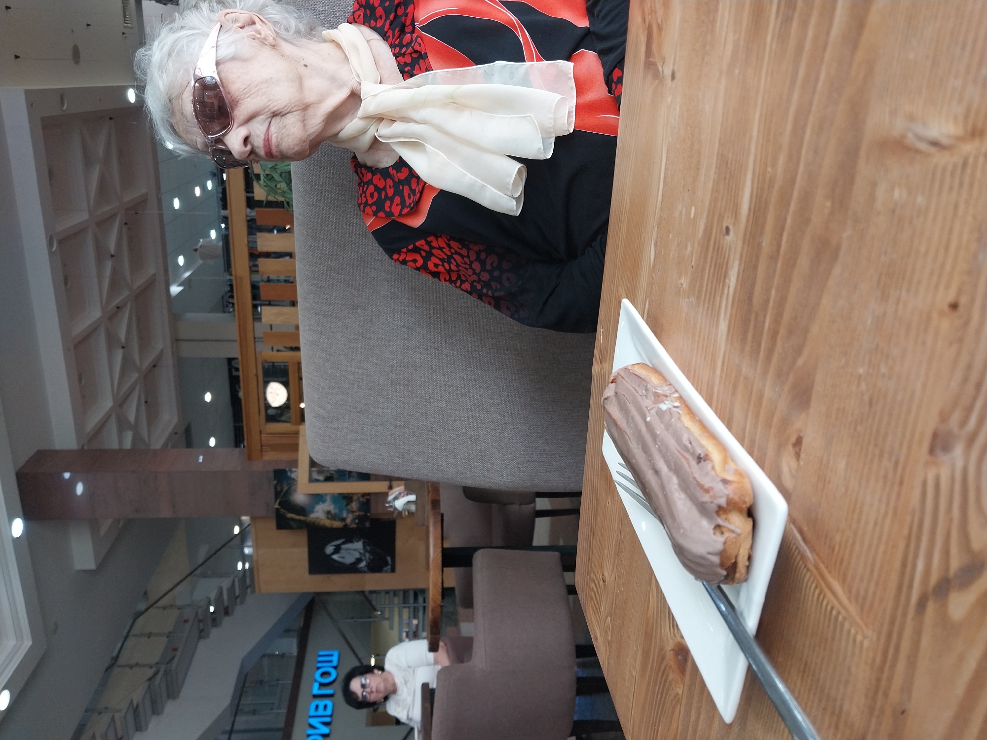 An elderly woman with white hair and sunglasses is sitting at a table in a cafe. She is wearing a black and red patterned top and a cream-colored scarf. In front of her on the table is a white plate with an eclair and a fork. In the background, another woman is sitting at a table, and there is a staircase and a sign with Cyrillic text that reads "РИВ ГОШ". The cafe appears to be located in a shopping center.