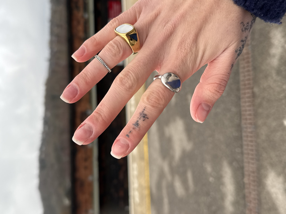 A hand is displayed with three rings. The index finger has a gold signet ring with a white stone. The middle finger has a thin silver band. The ring finger has a silver ring with a blue stone. The hand is pale with visible veins. There is a tattoo of vertical lines on the side of the middle finger, and another tattoo on the side of the hand, near the wrist. The nails are neatly shaped and have a natural look.