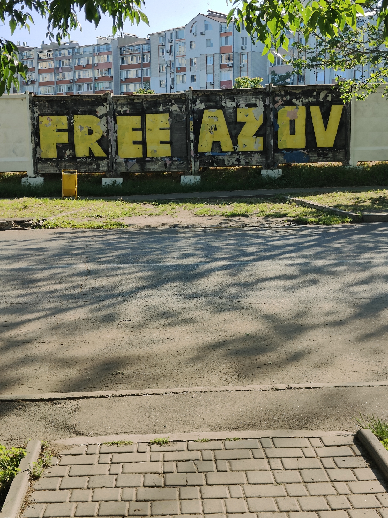 This outdoor image captures an urban street scene in Chornomorsk, Ukraine, on a bright, sunny day. In the immediate foreground, a grey paved sidewalk transitions into an asphalt road. The road surface is extensively covered in dappled shadows cast by overhead tree leaves, indicating a clear sky and either mid-morning or afternoon sun.
Beyond the road and a strip of green grass, a long, multi-panel fence or barrier dominates the midground. This fence is dark-colored, possibly black or dark blue, and features large, yellow-painted graffiti spelling out "FREE AZOV." The yellow paint shows signs of wear, fading, and chipping across the letters. A yellow, cylindrical trash bin is positioned on the grass directly in front of the "FREE" section of the fence.
In the background, several multi-story residential buildings are visible, typical of apartment complexes with numerous windows, balconies, and external air conditioning units. These buildings appear to be of a light color, possibly white or light grey, with some darker architectural elements. Green leaves from trees partially obscure the top corners of the frame. No individuals are present in the scene. The image prominently features the political message "FREE AZOV," displayed on a worn urban structure, against a backdrop of everyday residential life.