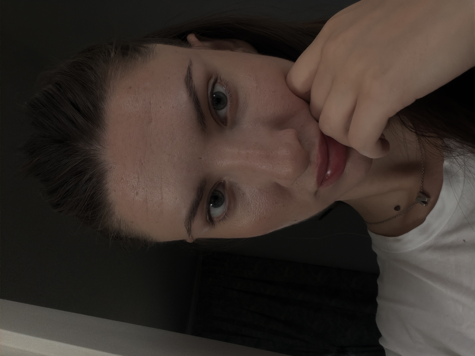 A young woman is captured in a close-up, head-and-shoulders shot, looking directly at the camera with a slightly pensive or curious expression. Her right hand is raised, with her fingers gently touching her chin and cheek, creating a sense of introspection. She is wearing a simple white t-shirt and a delicate silver necklace with an 'N' pendant.
The lighting in the image is subdued, with shadows dominating the background and a soft light illuminating her face from the front. This creates a moody and intimate atmosphere. Her hair is pulled back, revealing her forehead and the contours of her face. The background is dark and indistinct, with hints of fabric suggesting curtains or a similar soft furnishing, and a vertical element on the left that could be part of a doorway or frame.
There is no information or visual cues within the image that would place the scene in Sharm El Sheikh, Egypt, or any specific urban location. The setting appears to be indoors, in a dimly lit room. The focus is entirely on the woman and her immediate presentation.