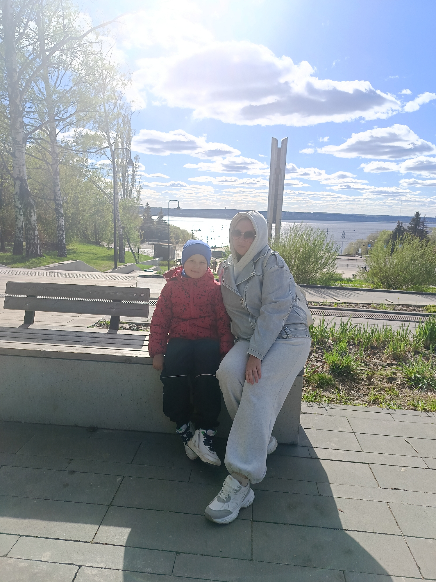 A medium, outdoor shot depicts a woman and a young boy sitting on a concrete bench. The woman is wearing a light gray hooded sweatshirt, matching sweatpants, and white athletic shoes. She is also wearing sunglasses. The boy is wearing a blue hat, a red puffy jacket, black pants, and white athletic shoes. They appear to be posing for the picture.
In the background is a view of a river or large body of water under a bright, partly cloudy sky. Trees and vegetation are visible on the opposite bank. A concrete monument can be seen to the right of the woman. The image is taken in a park or public space with a tiled walkway and green areas. Based on the architectural features and the monument in the background, this is likely a location in the city of Izhevsk, Russia.