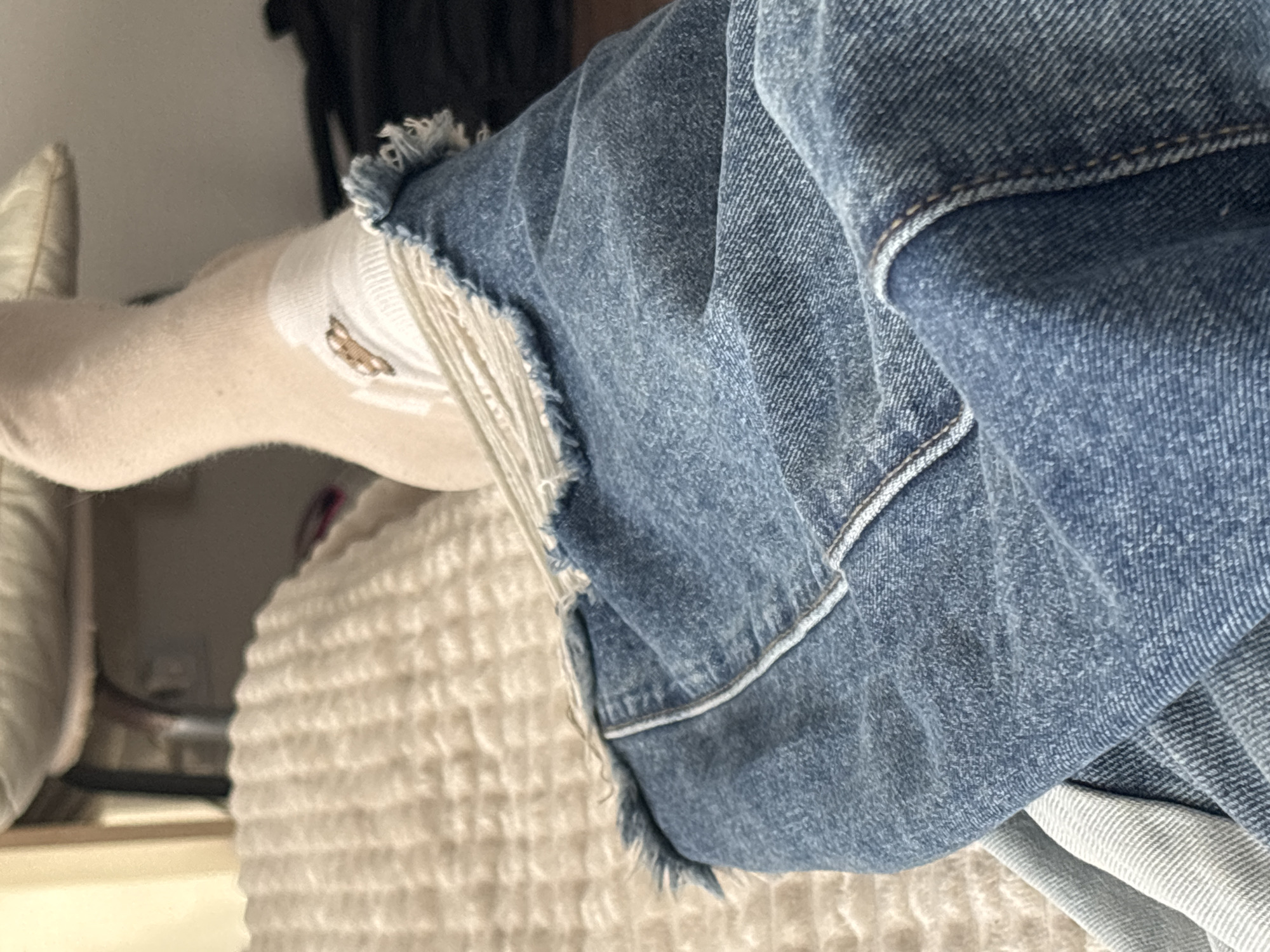 A close-up shot depicts a person's lower leg and the bottom of a pair of light-wash denim shorts. The leg is covered with a beige sock that has a small, brown graphic on the ankle. The denim shorts feature a frayed hem. The subject is resting on a textured, beige blanket or cushion. The background is slightly blurred, but a beige pillow and a dark, indistinct object are visible. The location, Vsevolozhsk, Russia is not visually evident in the image.