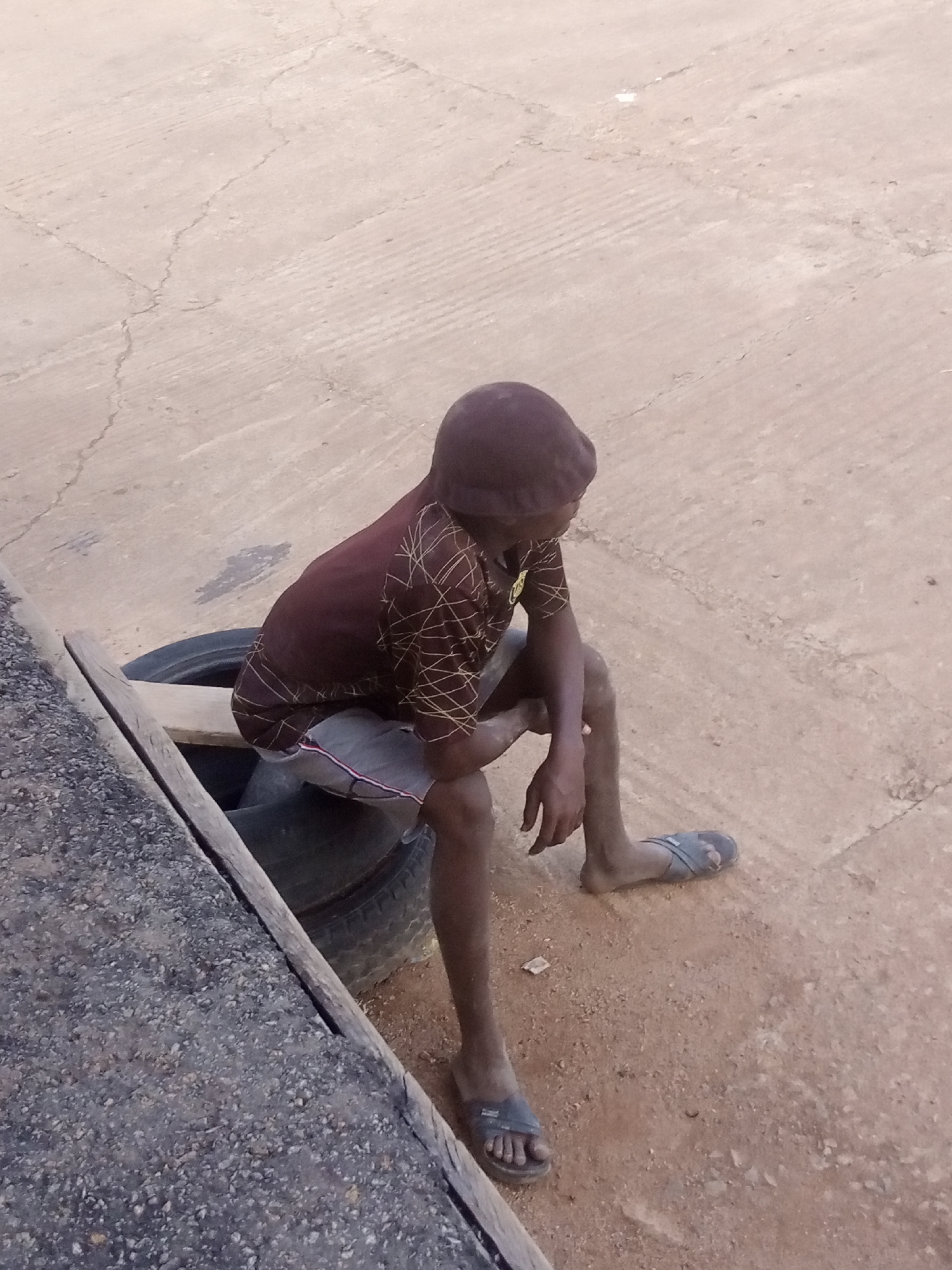 A young person wearing a maroon shirt with a yellow geometric pattern and grey shorts is sitting on stacked tires. The person is wearing blue sandals and a maroon cap. The scene is outdoors on a paved surface with cracks. A wooden plank is visible on the left side of the image.