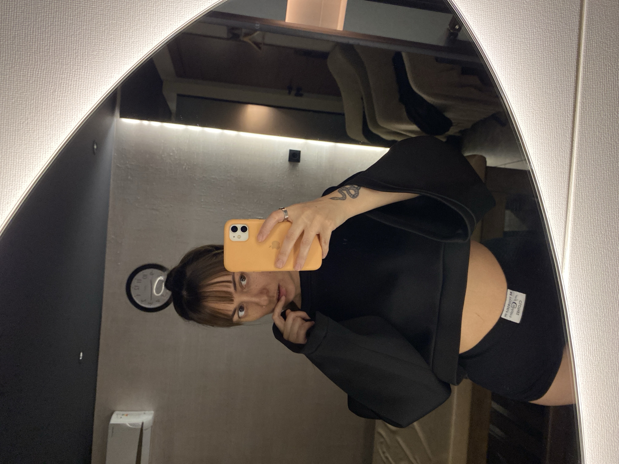 A woman is taking a selfie in a mirror. She has brown hair with bangs and is wearing a black crop top and black shorts. She is holding an orange smartphone. A clock hangs on the wall behind her. The location is likely a clothing store fitting room, based on the mirror and the reflected clothing racks. The city and country are indicated by the location of the clothing company on the shorts, "Cluuby," which operates within the Russian Federation and specifically the area of Votkinsk.