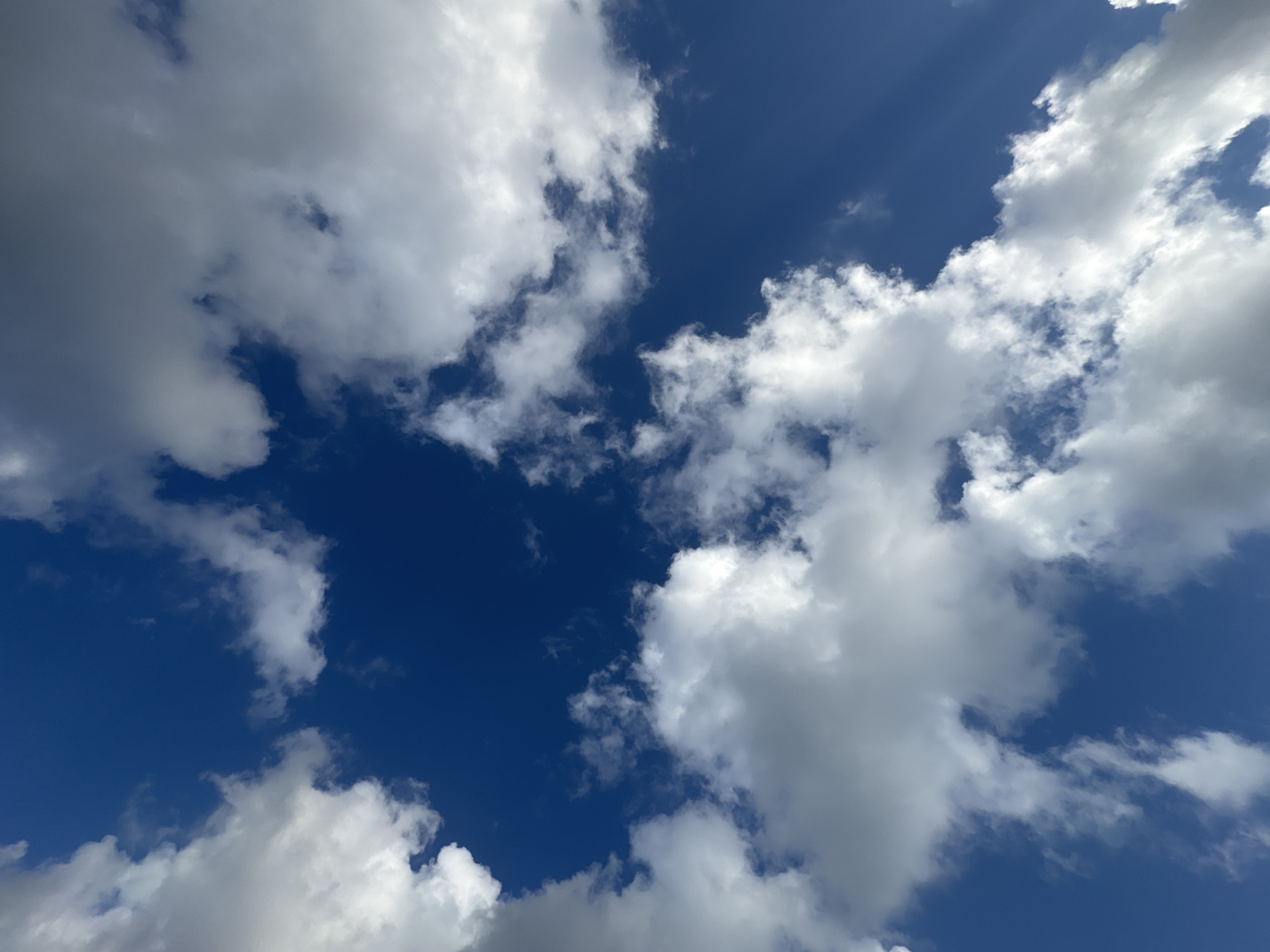 A low-angle shot captures a bright blue sky filled with scattered white and gray clouds. The clouds vary in density, with some appearing wispy and others more solid. Shafts of sunlight are visible breaking through some of the denser cloud formations, creating a dynamic and illuminated effect. The image appears to be taken outdoors in Kuta Selatan, Indonesia. There are no people or man-made objects visible in the frame.