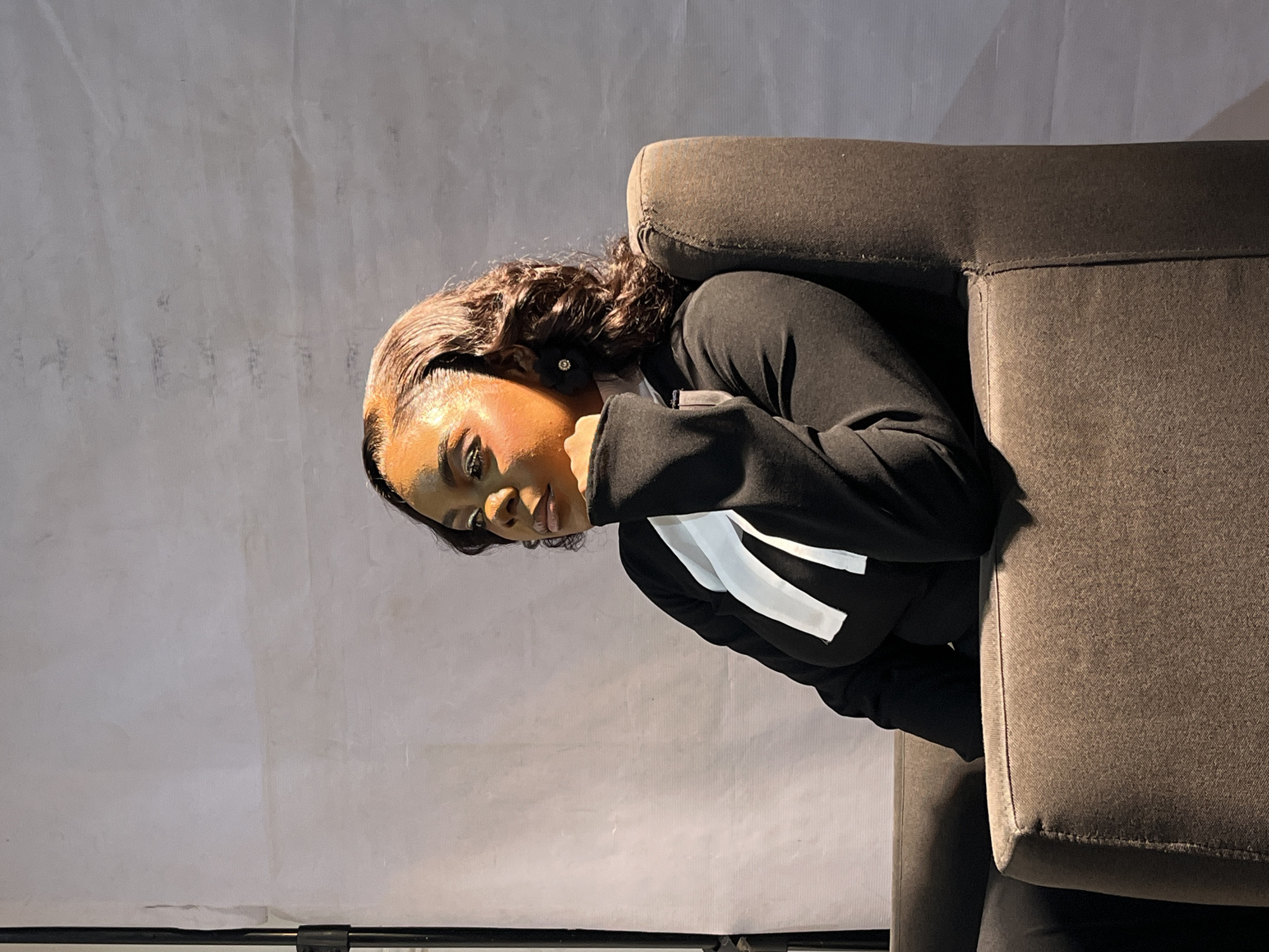 A woman of color with dark curly hair and noticeable makeup, including glittery eyeshadow and a defined contour, is seated in a dark upholstered armchair. She is wearing a black long-sleeved top with white lapel-like accents. Her left hand is resting on her chin, and she is looking off to her left. The background is a plain, light-colored wall with some subtle texture. A lighting stand is visible on the left edge of the frame. The scene appears to be indoors, possibly in a studio setting, with professional lighting casting strong highlights and shadows on the subject.