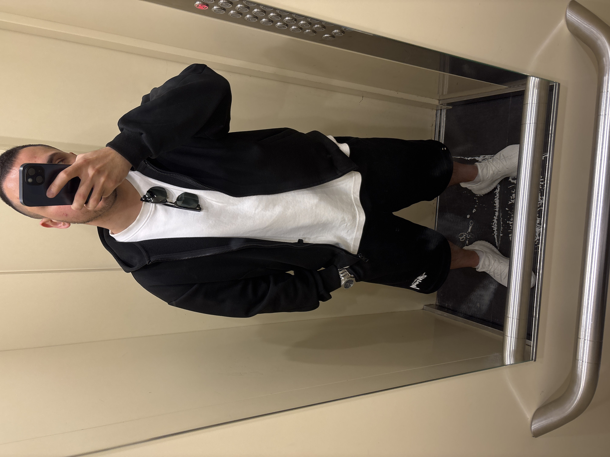 A male individual is featured taking a mirror selfie inside an elevator. He is holding a black smartphone with an exposed Apple logo on the back, partially obscuring his face. He is dressed in a black zip-up hoodie over a white t-shirt, black shorts, and white sneakers. Sunglasses are hooked onto the collar of his t-shirt. A watch with a metallic strap is visible on his left wrist.
The activity depicted is a mirror selfie being taken by the person. His reflection fills most of the frame, which is rotated 90 degrees counter-clockwise.
Notable details include the setting, which is clearly an elevator with mirrored walls, a control panel featuring numerous silver buttons, and a chrome handrail. The elevator floor appears to be dark and slightly worn. On one red button on the control panel, the letter "S" is visible. A white, stylized logo, possibly "Stussy," is printed on the left leg of his shorts. The lighting suggests an indoor, artificially lit environment. The person's expression is not fully discernible due to the phone position.
