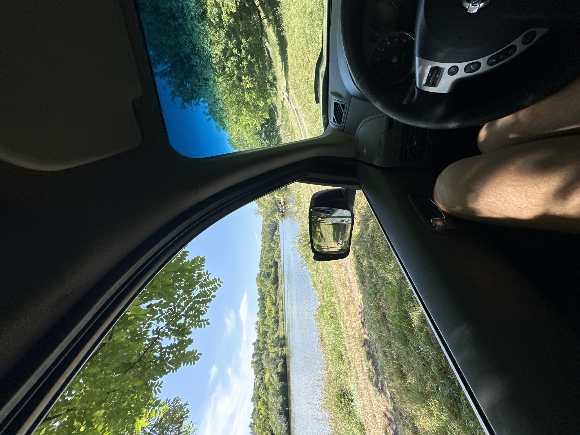 The image shows a first-person view from inside a car, parked near a body of water and surrounded by greenery. The perspective is looking out the driver's side window and through the windshield. The environment appears to be a natural setting, possibly a rural area or park. The water is calm and reflects the sky, with trees and vegetation lining the opposite bank. In the foreground, inside the car, the steering wheel and dashboard are visible. A person's legs are also visible, indicating the presence of an occupant in the driver's seat. The weather appears to be sunny with a clear blue sky and some scattered clouds. The location is identified as Makiv, Ukraine.