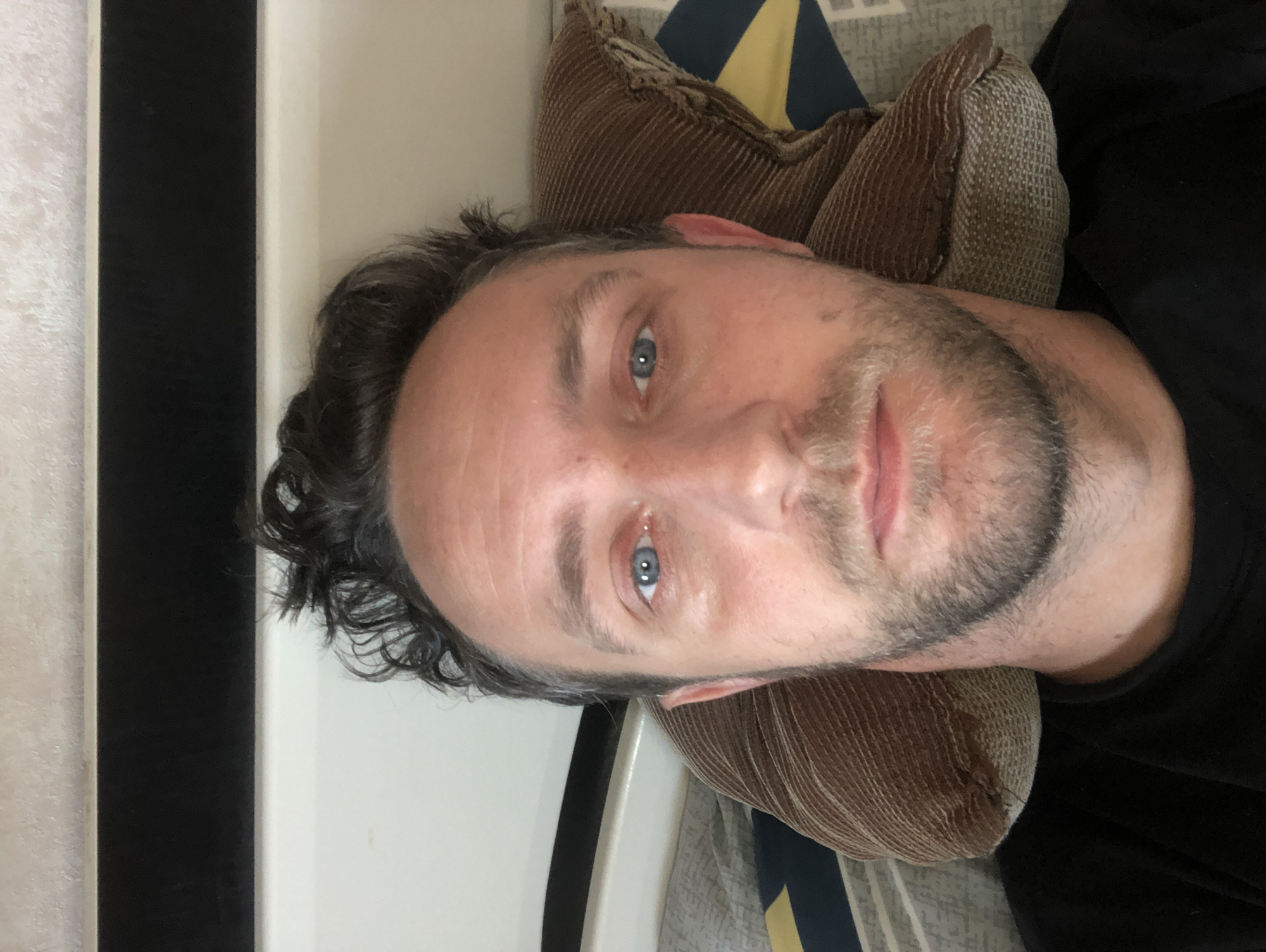 The image is a selfie taken from a reclined position, looking upwards. A man with short, dark, curly hair and a beard is the primary subject. He has blue eyes and appears to be indoors, possibly in a bedroom. He is lying on a pillow with a corduroy texture, which is placed on a bed with a patterned duvet. A dark, horizontal object is visible at the top of the image, suggesting a headboard or a shelf. The lighting is natural, coming from above and slightly to the left. The background appears to be a wall with a textured surface. There is no information in the image that suggests a specific location in Baranavichy, Belarus.