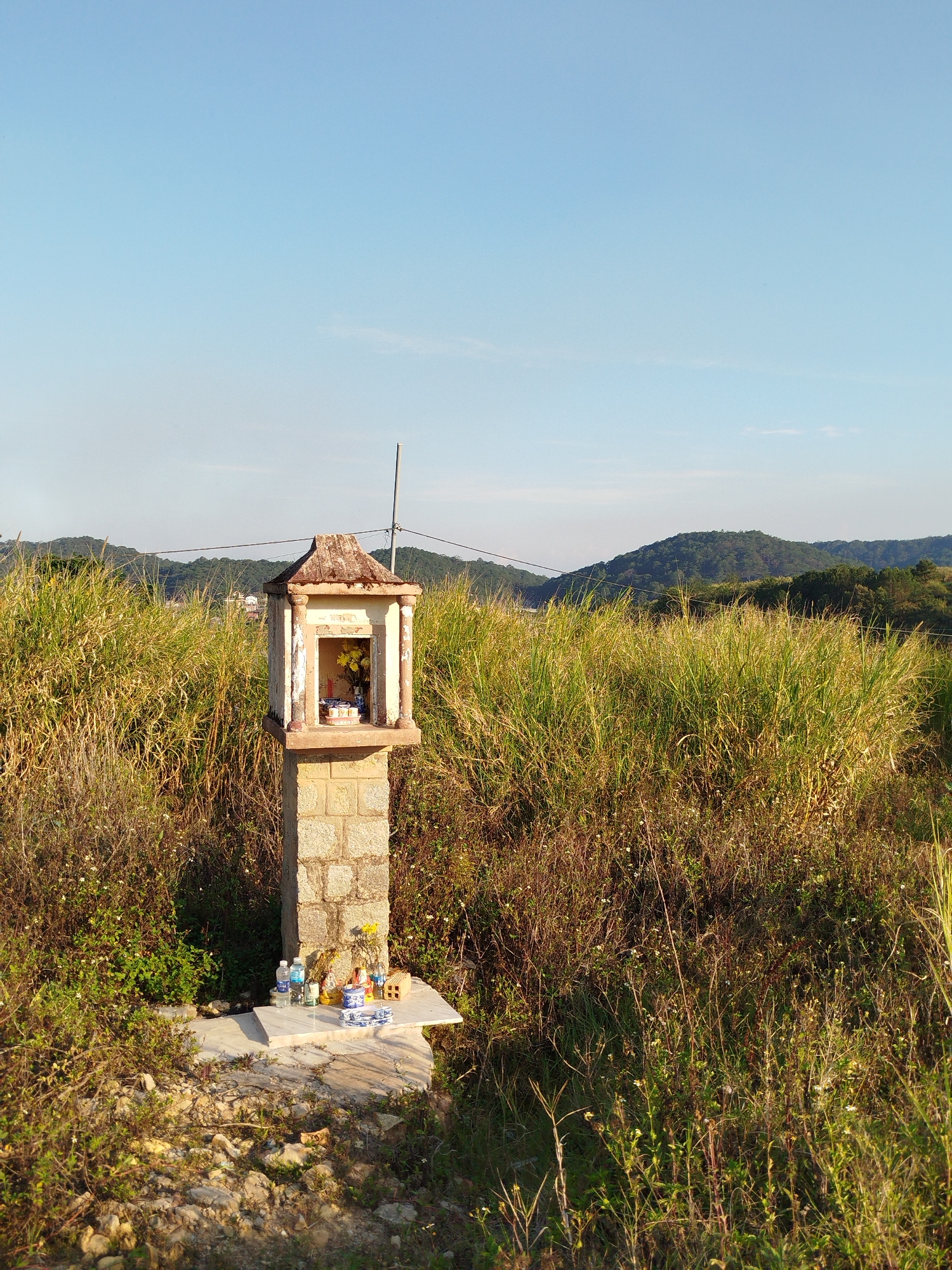 A small, weathered shrine stands amidst tall grasses and low scrub vegetation in Da Lat, Vietnam. The shrine is made of stone blocks and has a peaked roof. Inside the shrine are offerings of flowers and incense. At the base of the shrine are additional offerings including bottled water and ceramic dishes. The background features rolling hills partially covered in vegetation and a clear blue sky. An electrical pole and wires are visible behind the shrine. The ground around the shrine is rocky and uneven. The image is taken during daylight, with sunlight casting shadows on the foreground vegetation.