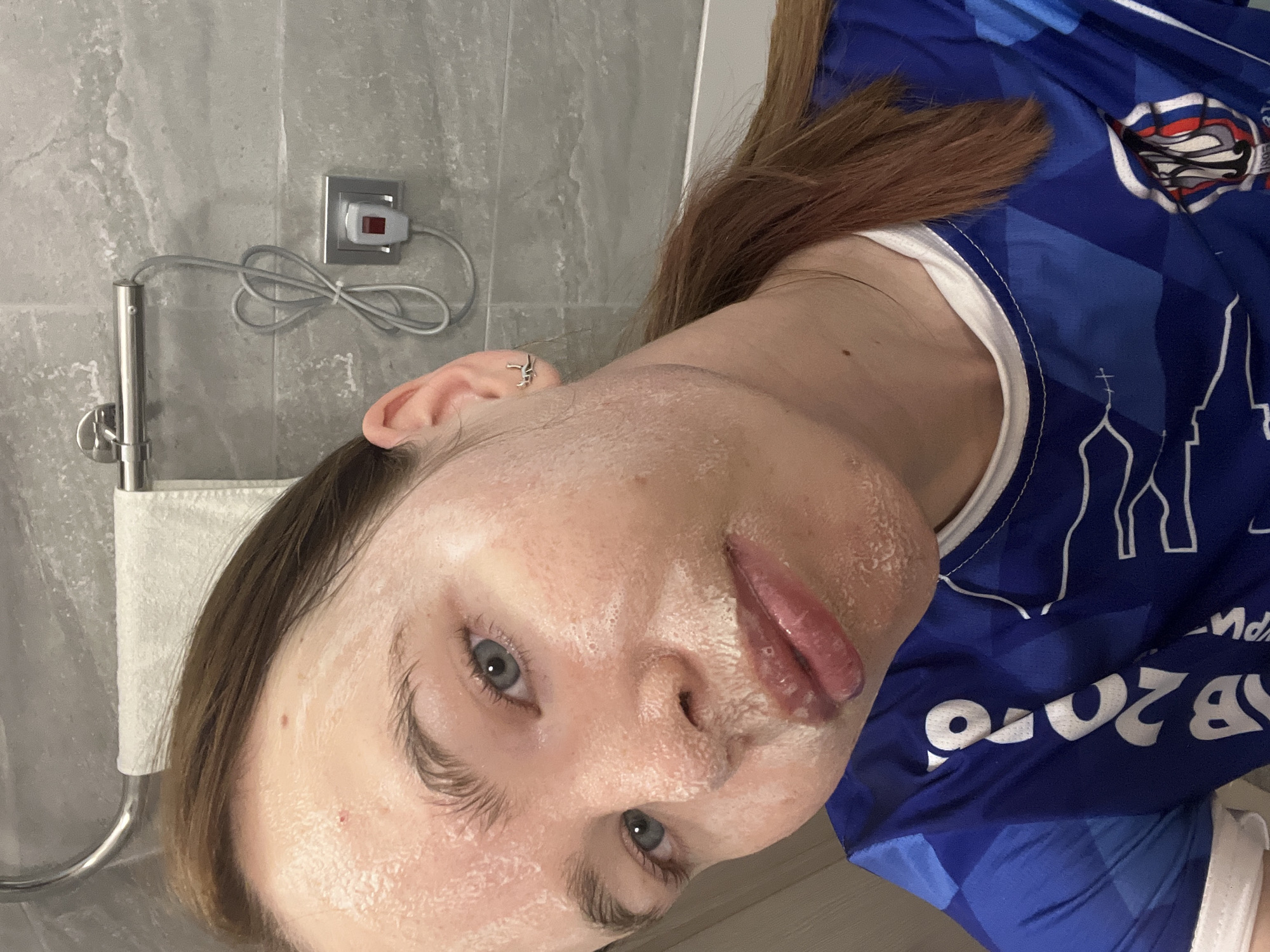 A person with fair skin and light brown hair is in a bathroom. The person has a white face mask applied to their face. They are wearing a blue sports jersey with white accents and lettering. The jersey also has a graphic of a building with a spire, and text that appears to read "2016." The person's lips appear glossy.
The background includes a light gray, textured wall, a chrome towel rack with a white towel, and an electrical outlet with a cord. The photo appears to be taken in a bathroom setting. I cannot determine the city of Perm, Russia from the image.