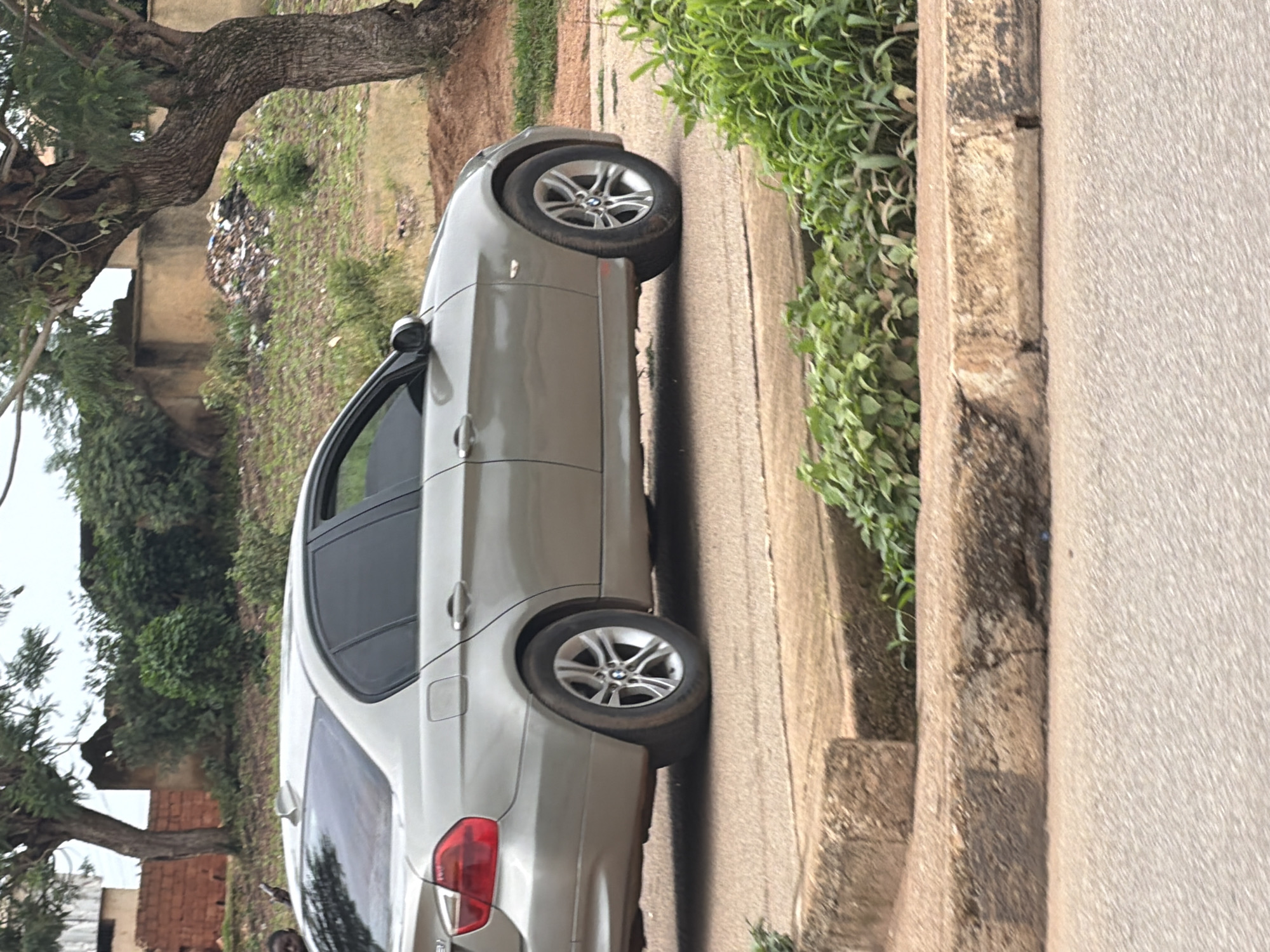 A silver BMW sedan is parked on a paved road in Bukuru, Nigeria. The car is viewed from the side, showing its passenger side. The vehicle has tinted windows and multi-spoke silver alloy wheels. In the background, there are trees, some buildings constructed with brick and concrete, and a pile of debris and garbage. The foreground shows a concrete curb and more greenery. A person's head and shoulder are visible on the far left of the image, near the rear of the car.
