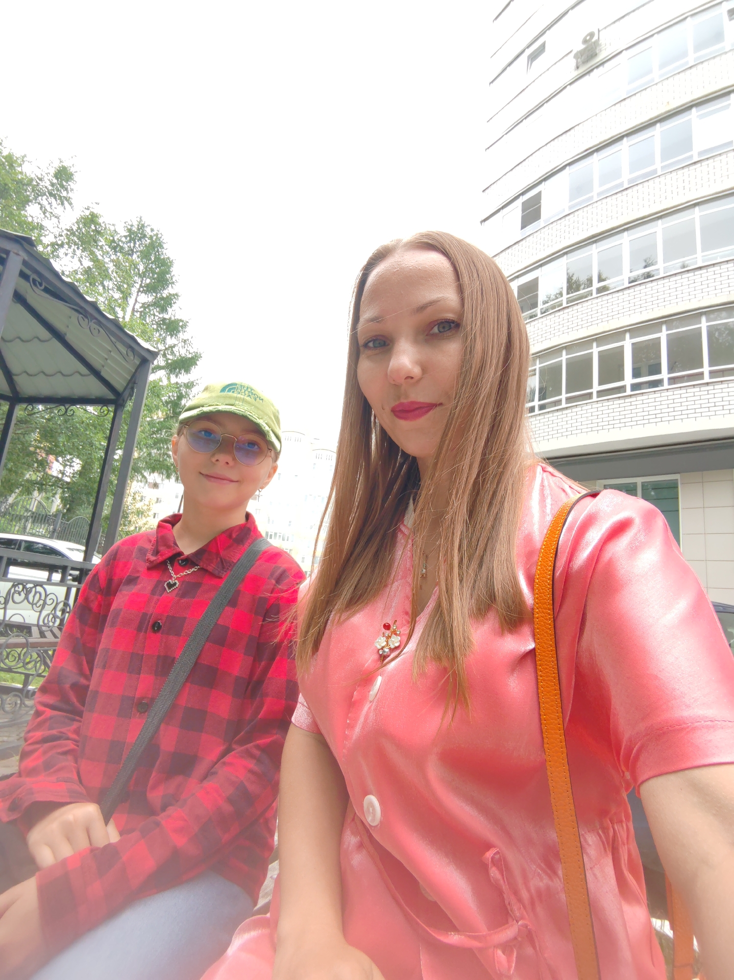 A young person wearing a red and black plaid shirt, blue jeans, glasses, and a green cap is sitting on a bench next to an adult woman wearing a shiny pink dress. The two are outdoors with a building in the background. It appears to be daytime and the sky is overcast. The woman is taking a selfie with the young person, and they are both looking at the camera. This scene is likely in the city of Syktyvkar, Russia, given that this is a popular spot for locals and tourists alike. The building in the background is a residential building, which is common in Syktyvkar. The trees and greenery suggest that this is a park or a residential area. The bench the two are sitting on appears to be a park bench, further supporting the idea that they are in a public outdoor space.