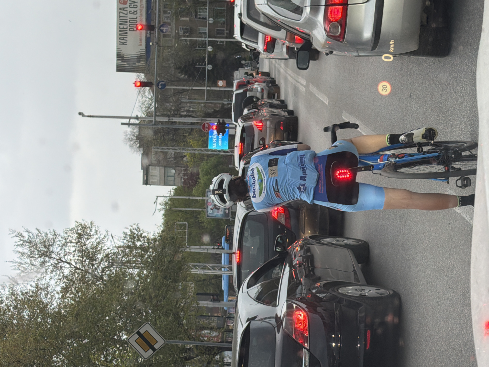 A vertically oriented, eye-level photograph captures a street scene dominated by a traffic jam under an overcast sky. The primary subject is a male cyclist, wearing a light blue and dark blue racing kit with white accents, a white helmet, and cycling shoes, standing beside his blue road bike. He is positioned among several cars, facing forward in the queue.
The street is densely packed with vehicles, with brake lights visible on many cars, indicating they are stopped. Traffic lights in the distance and closer to the cyclist are red, reinforcing the stopped traffic. In the background, there are buildings and trees with sparse foliage, suggesting spring or autumn.
Notable details include:
* **Cyclist's gear:** His helmet has "poc" written on it. His jersey features Cyrillic text, including "Бачково" (Bachkovo) and "Армеец" (Armeec). His bicycle frame has "GAS" visible. A bright red rear light is active on his bike.
* **Road markings/signs:** A "30" km/h speed limit is painted on the road, alongside an overlay of "0 km/h," likely from the camera's heads-up display. A diamond-shaped "priority road" sign is visible near a tree. A "STOP" sign is present near a billboard.
* **Billboards/Text:** A large billboard advertises "KAMENITZA POOL & GYM" with "kamenitzapoolandgym" below it. A smaller blue sign has Cyrillic text "ЗА РЕКЛАМА" (For Advertising) and a phone number "0887 98".
The scene depicts a common urban scenario of a cyclist navigating congested city traffic.
