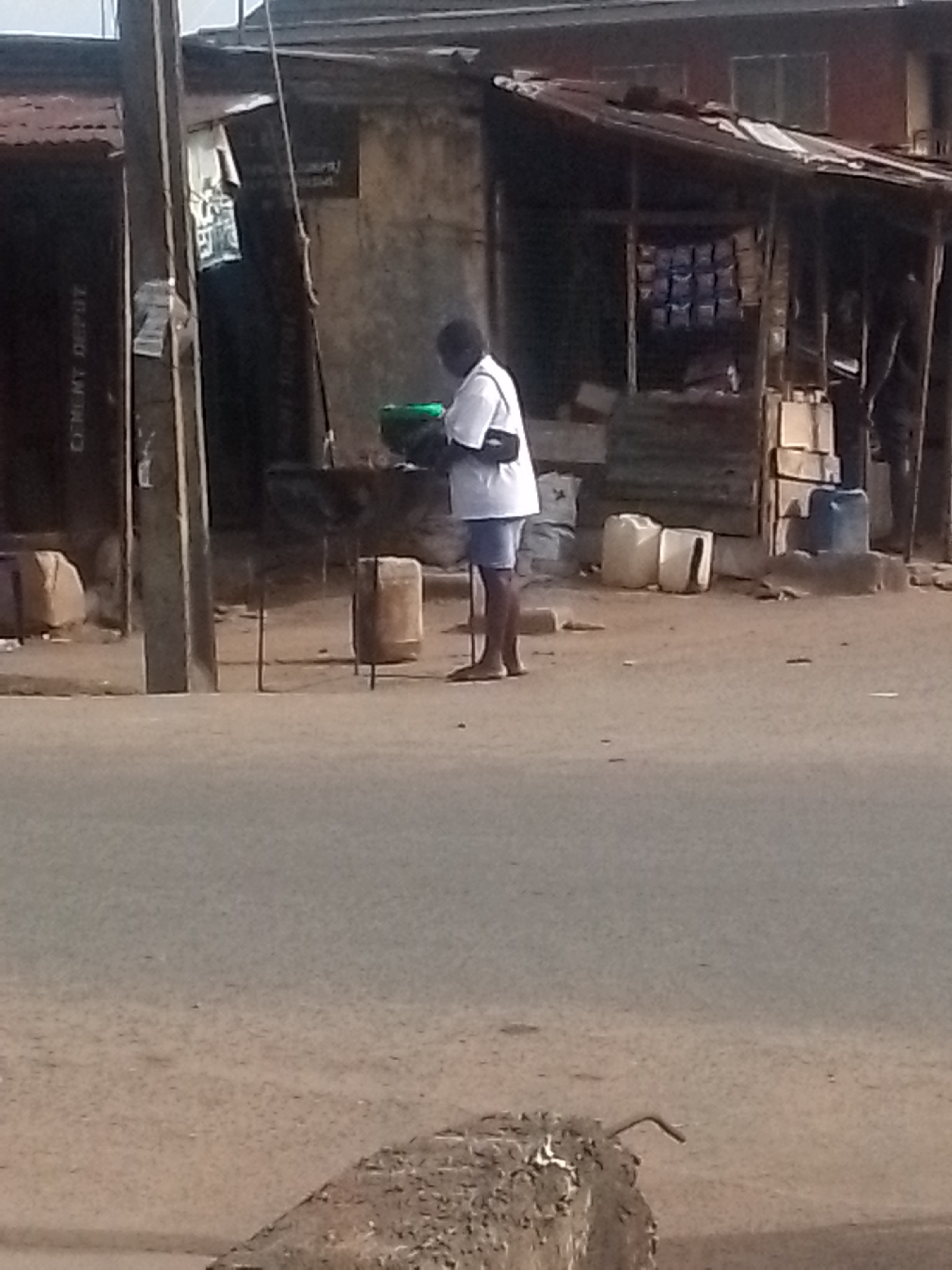 The image depicts an outdoor street scene in an urban area, potentially a commercial district. A man wearing a white shirt and dark shorts is standing in the street, carrying a green container. He has a strap or bag across his shoulder. To the left is a building with a sign that reads "CEMENT DEPOT". There are makeshift structures or stalls with goods on display. The road surface appears to be unpaved or roughly surfaced. There are utility poles and various containers visible in the background. The overall setting suggests a lower-income or developing urban environment.