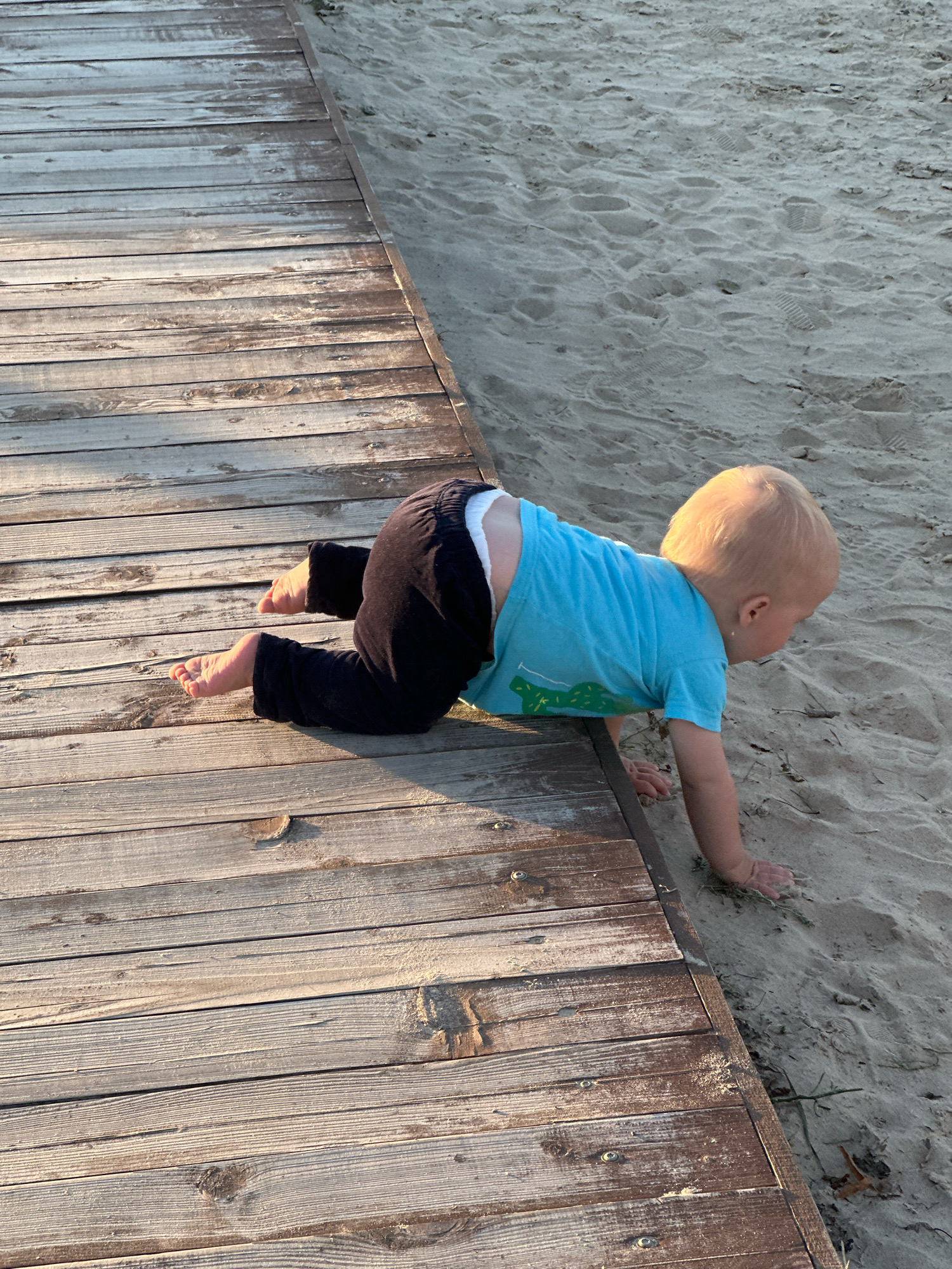 A young child is crawling on a wooden boardwalk next to a sandy beach. The child is wearing a blue t-shirt with a green graphic and dark pants. The child's head is turned to the right, looking towards the sand. The boardwalk is made of weathered wooden planks and is adjacent to a sandy area with footprints. The scene appears to be outdoors during daylight.