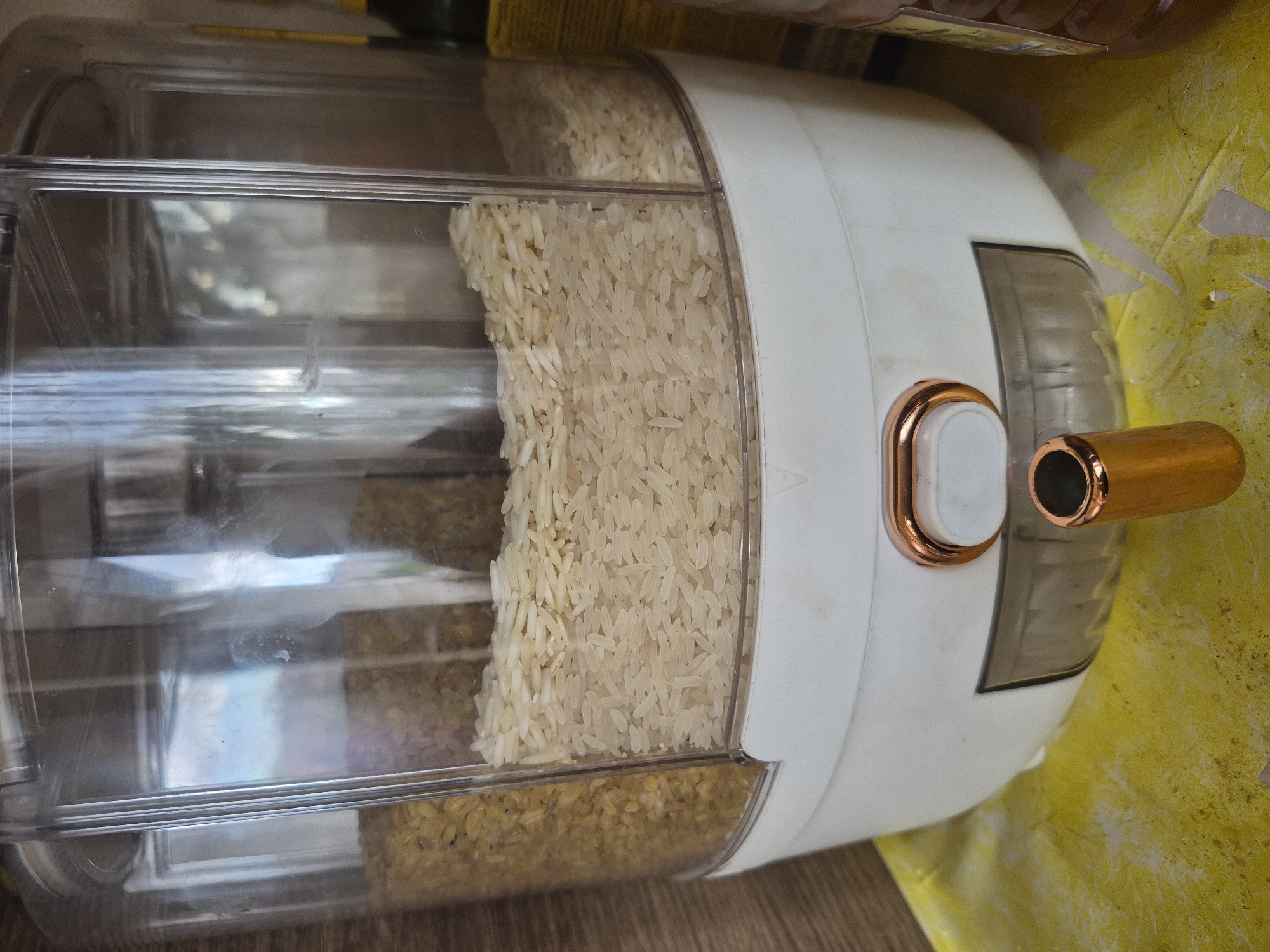 The image displays a close-up, slightly tilted view of a modern grain dispenser in an indoor setting. The dispenser is primarily made of clear, rectangular plastic, allowing its contents to be visible. Its top section is white, featuring a rose gold-colored trim around a white push-button or lever mechanism, and a matching rose gold-colored cylindrical spout or handle on its right side.
The dispenser is filled with two different types of grains. The upper compartment contains a substantial amount of light beige, long-grain rice. Below it, in the lower compartment, are smaller, yellowish-brown grains, possibly split peas or lentils.
The dispenser rests on a brown wooden surface, which appears to be a table or shelf. In the background, particularly to the right and behind the dispenser, there is a crinkled, textured yellow material, resembling a crumpled bag or packaging, showing some torn edges. Blurred objects, possibly other containers, are faintly visible in the far background.
The clear plastic surface of the dispenser exhibits some reflections and minor smudges. No people are present in the image, and no discernible text is visible. The scene suggests a static moment, focusing on a household item used for storing dry goods, located in Yerevan, Armenia.