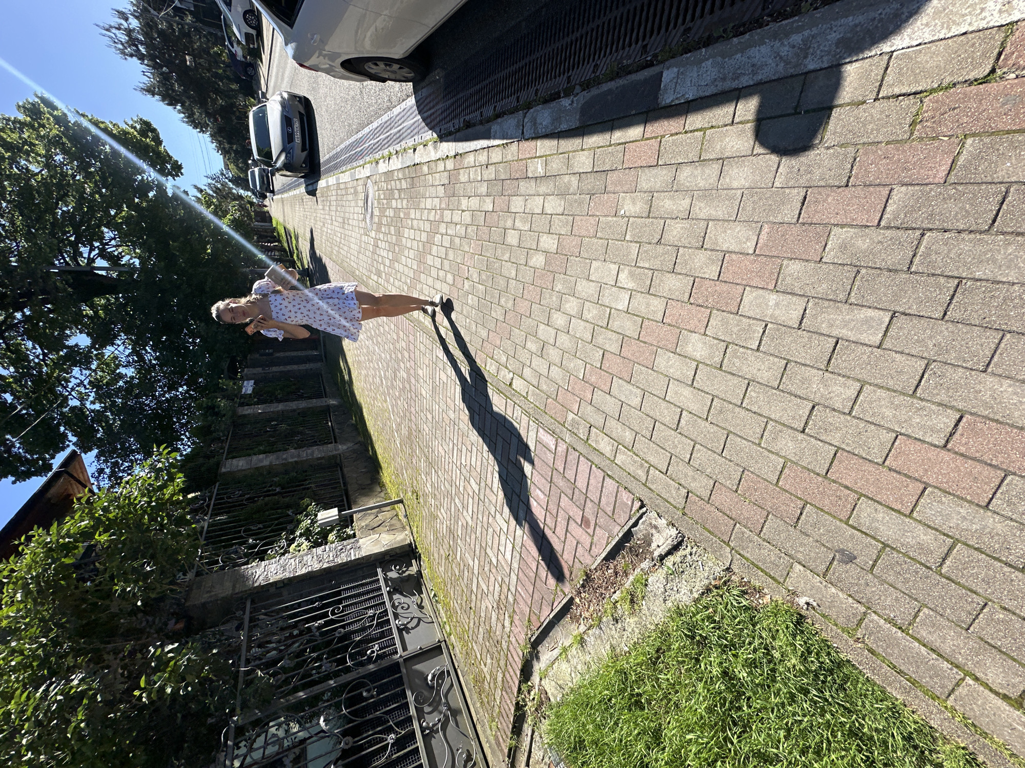 The image captures a sunlit street scene in Krasnaya Polyana, Russia, viewed from a slightly elevated angle. A young woman is the central subject, walking or standing on a patterned sidewalk. She has blonde hair and is wearing a white short-sleeved dress adorned with small red floral patterns. Her long, distinct shadow stretches ahead of her on the pavement, indicating bright sunlight, likely mid-morning or late afternoon.
The sidewalk is composed of rectangular paving stones in alternating shades of light grey and reddish-brown. To the woman's right, the street is visible with several parked cars. A silver sedan in the mid-ground displays a license plate with "X169PO 94". Beyond the cars, large green trees line the street under a clear blue sky, with a strong sun flare visible in the upper left.
To the woman's left, a dark, ornate metal fence with a gate separates the sidewalk from an overgrown area with dense green foliage and parts of a building featuring balconies with similar metal railings and stone facade accents. Patches of bright green grass and some dry vegetation grow along the edges of the sidewalk. The scene conveys a bright, pleasant day.