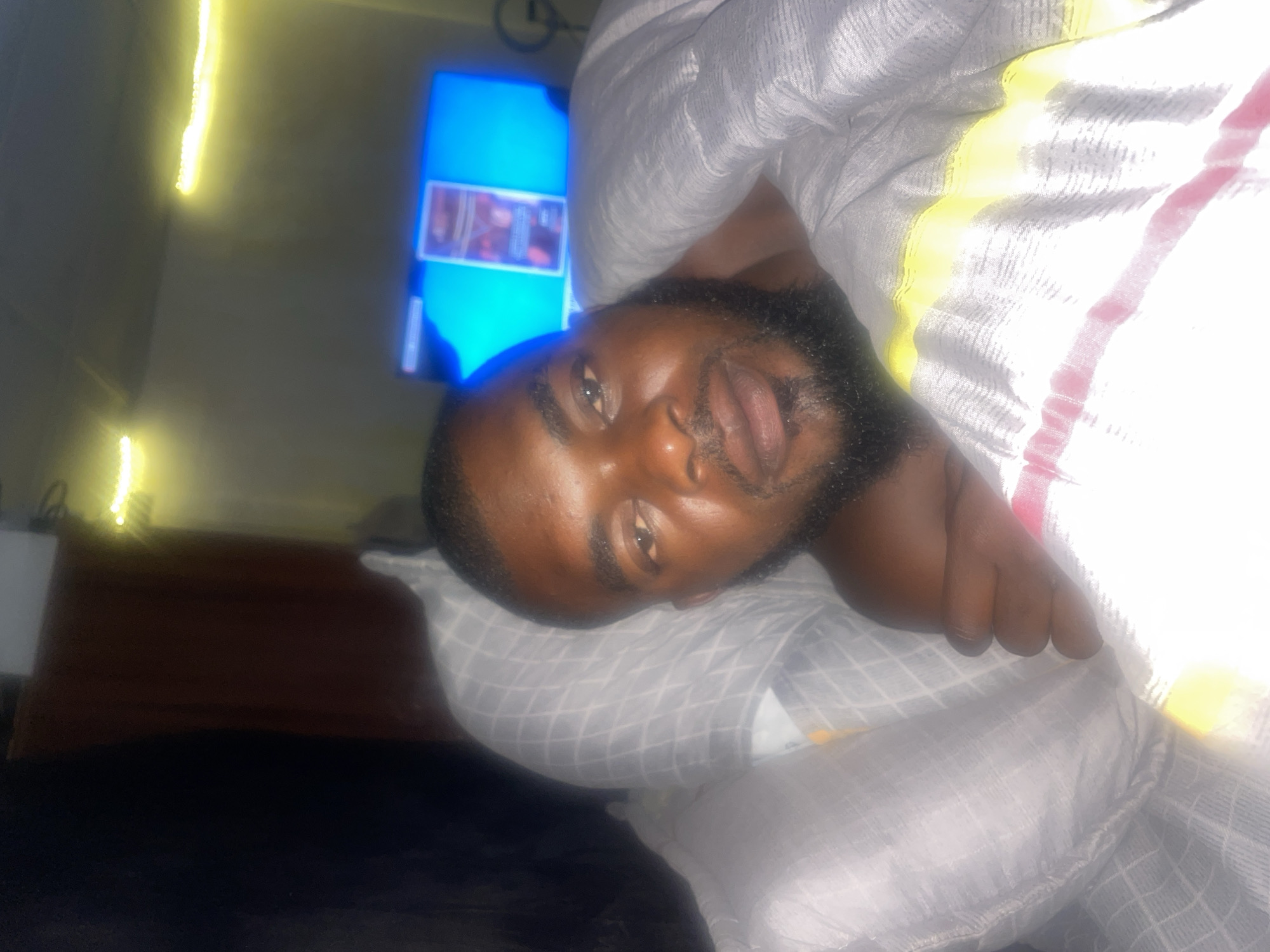 A man is lying in bed, appearing relaxed. He is under a patterned white blanket with yellow and red stripes. His head rests on a grey checkered pillow. A television screen is visible in the background, displaying images. The location is indicated as Jos, Nigeria. Additional details: a piece of furniture and some lights are visible, but the image is overall unclear.