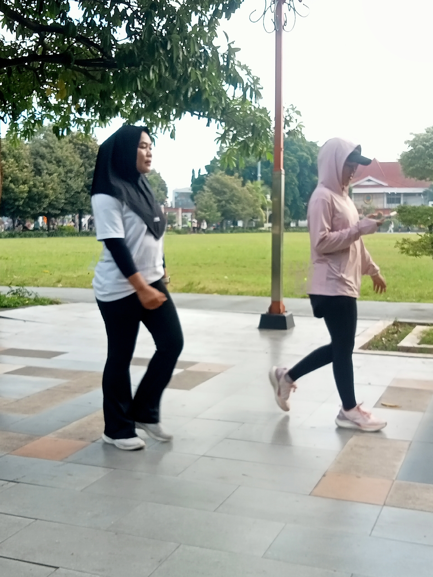 In the city of Kecamatan Pati, Indonesia, two women are captured walking on a paved pathway adjacent to a grassy area with trees. The woman on the left is wearing a black hijab, a white t-shirt with black sleeves, and black pants. The woman on the right is wearing a pink hooded jacket, a black baseball cap, and black leggings. Both are wearing athletic shoes. The pathway is composed of light-colored rectangular tiles with some darker and reddish-brown accents. In the background, there is a building with a red roof and a lamppost. The environment appears to be an outdoor public space, possibly a park or a promenade.