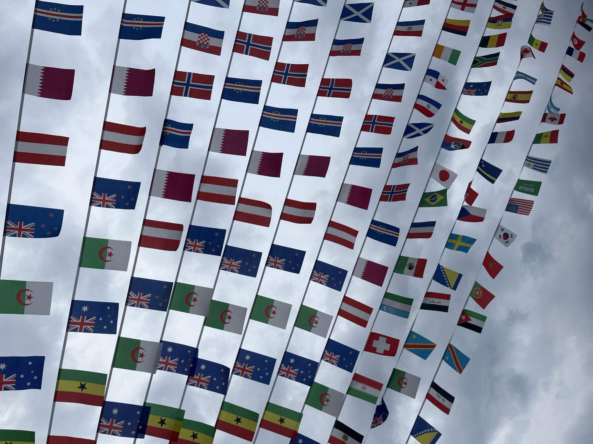 The image shows rows of various national flags suspended against a cloudy sky. The flags appear to be hung on lines stretching across the frame, creating a visual display. Flags from numerous countries are visible, including New Zealand, Australia, Algeria, Austria, Qatar, Croatia, Norway, Scotland, Cabo Verde, Japan, Brazil, Chile, Ghana, Switzerland, The Democratic Republic of Congo, South Korea, Tunisia, and many others. The location is suggested to be Domingo Arenas, Mexico. There are no people visible in the image. The dominant subjects are the flags and the sky. The interaction is between the flags and the wind causing them to wave. The environment is overcast, suggesting a cloudy day.