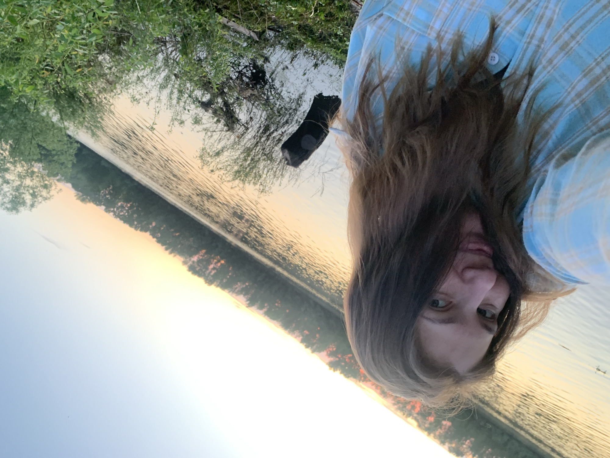The image depicts a selfie of a person wearing a light blue plaid shirt, with long brown hair partially obscuring their face. The background shows a body of water, likely a river or lake, reflecting the colors of a sunset or sunrise. The shoreline is visible with trees and vegetation. Part of a dark, indistinct object is near the water's edge. The location is potentially near the city of Chaikovsky, Russia, based on contextual information.