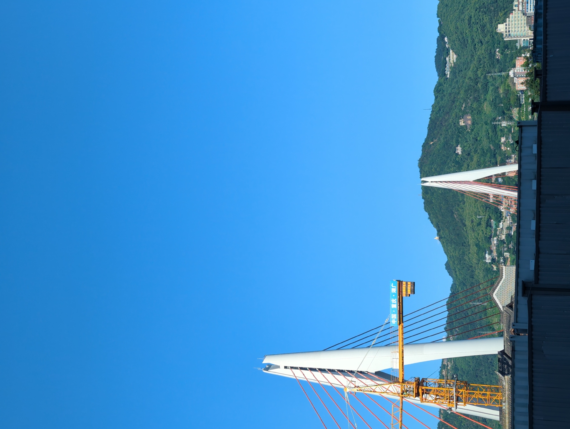The image shows a cityscape dominated by a clear blue sky and lush green mountains. In the foreground, a modern cable-stayed bridge pylon stands tall, with red cables extending from it. A yellow construction crane is attached to the pylon. Below the bridge, traditional tiled roofs are visible, interspersed with modern buildings. The mountainside is dotted with houses and some signage. On the mountain in the distance, large Chinese characters are visible. The overall scene depicts a blend of urban infrastructure, natural landscape, and residential areas, suggesting a location like Yuzhong District, Chongqing, China, known for its hilly terrain and bridges. There are no people visible in the image.