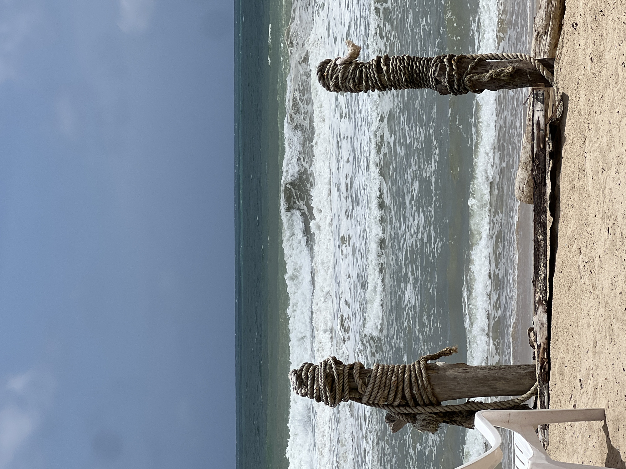 The image depicts a beach scene in Choeng Thale, Thailand, under an overcast sky. Two weathered wooden posts, heavily wrapped in thick rope, stand in the foreground. The post on the left is partially obscured by a white plastic chair positioned on the sandy beach. The posts are situated near the waterline, where the ocean exhibits moderate waves breaking. In the background, the sea transitions from a lighter, foamy green near the shore to a darker, greenish-blue further out, meeting the muted grey of the sky at the horizon. The sand on the beach appears light in color, with some scattered driftwood and debris visible around the base of the posts. The overall atmosphere is one of a calm, slightly turbulent coastal environment.