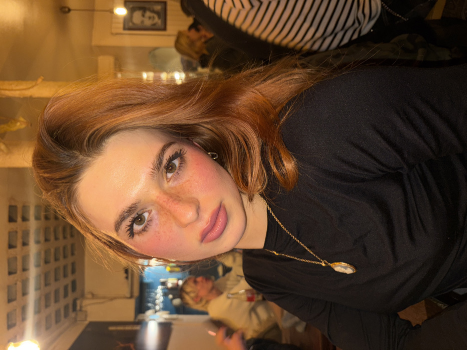 A young woman with auburn hair and freckles poses indoors. She's wearing a black long-sleeved shirt and a gold necklace with a cameo pendant. Her makeup includes noticeable blush. Other people are visible in the background, and there are shelves behind a table. The overall setting appears to be a bar or restaurant with ambient lighting. The image is slightly rotated.
