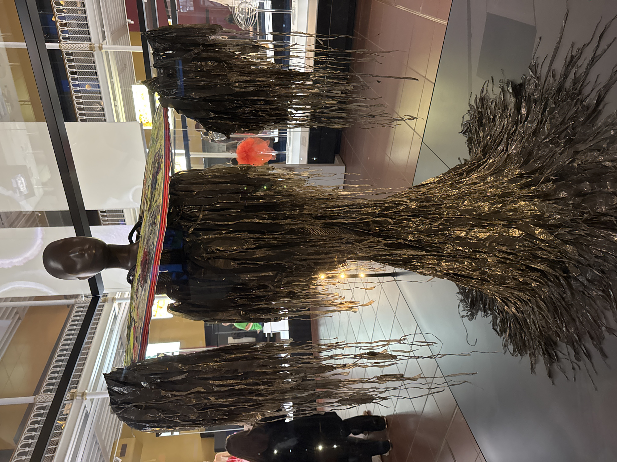 This image depicts an art installation in the National Museum of Scotland, located in the city of Edinburgh, United Kingdom. The central focus is a headless mannequin adorned with a garment constructed from dark, shredded material that resembles strips of plastic or fabric. This material hangs down from the shoulders and cascades to the floor, creating a voluminous, skirt-like effect. The mannequin is wearing a flat, rectangular headdress or accessory with a colorful, patterned surface. In the background, museum visitors are visible, some observing the display. The environment appears to be a museum hall with tiled flooring and architectural elements like railings and display cases.