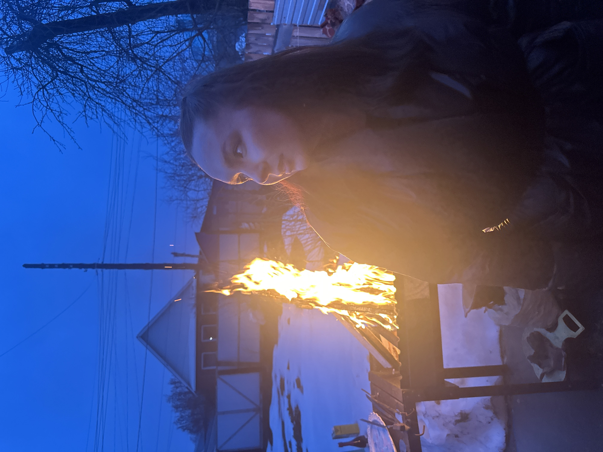 The image captures a woman standing next to a fire contained in a dark metal structure, possibly a grill or fire pit. The flames are bright and prominent, casting a warm glow on her face. She appears to be gazing at the fire. The surrounding environment shows signs of a cold season, with patches of snow on the ground. In the background, there is a house and a utility pole against a blue dusk sky. It is plausible that this image was taken in a residential area, but specific location details within the city of Chaikovsky, Russia cannot be determined from the image.