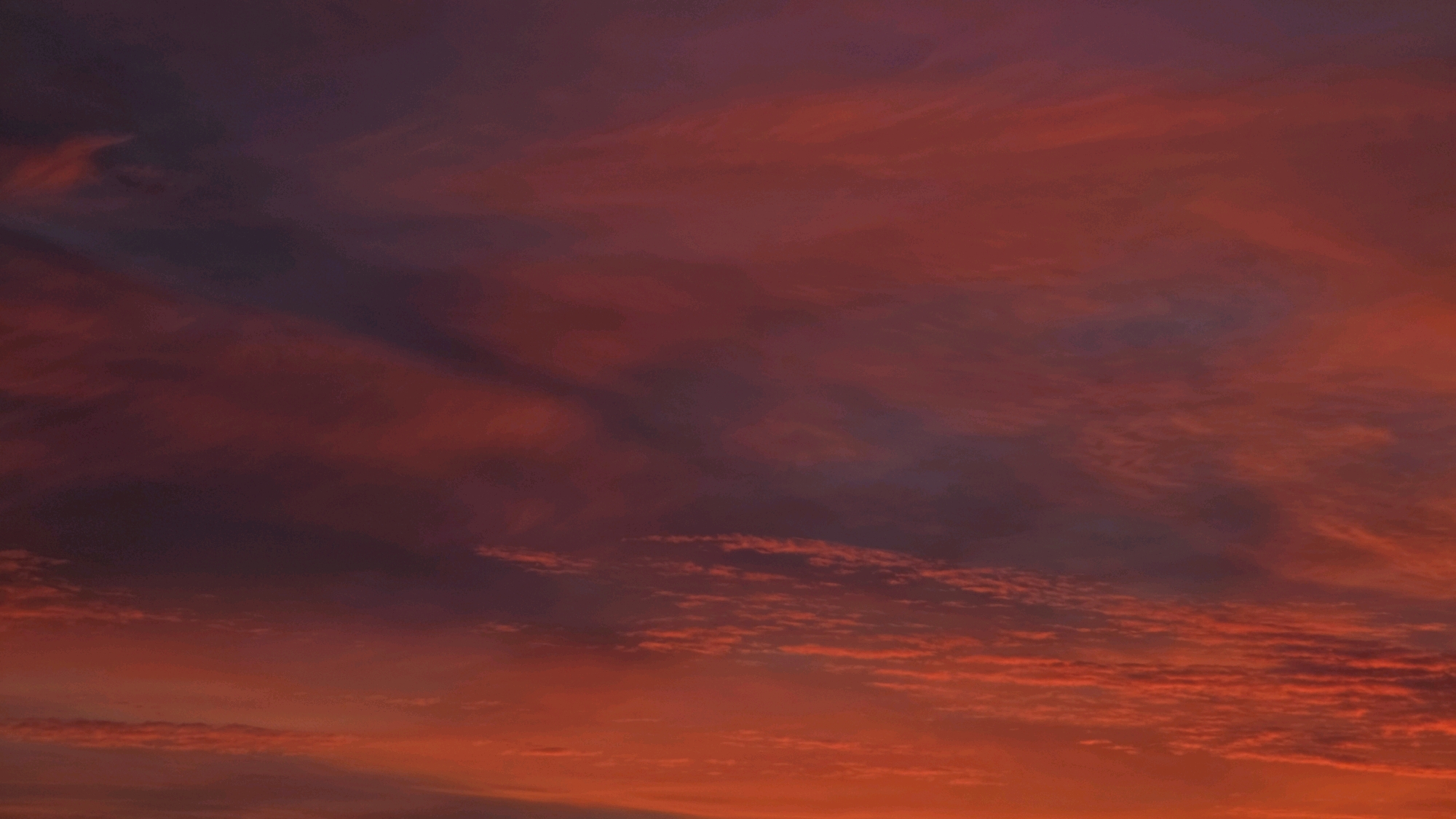 The image depicts a dramatic sunset over the city of Bad Kreuznach, Germany. The sky is filled with vibrant hues of orange, red, and purple, with streaks of clouds creating a textured, painterly effect. The lower portion of the image shows a hint of the cityscape, with faint outlines of buildings visible beneath the fiery sky. There are no people or discernible objects in the foreground or midground. The focus is entirely on the atmospheric display of the sunset.