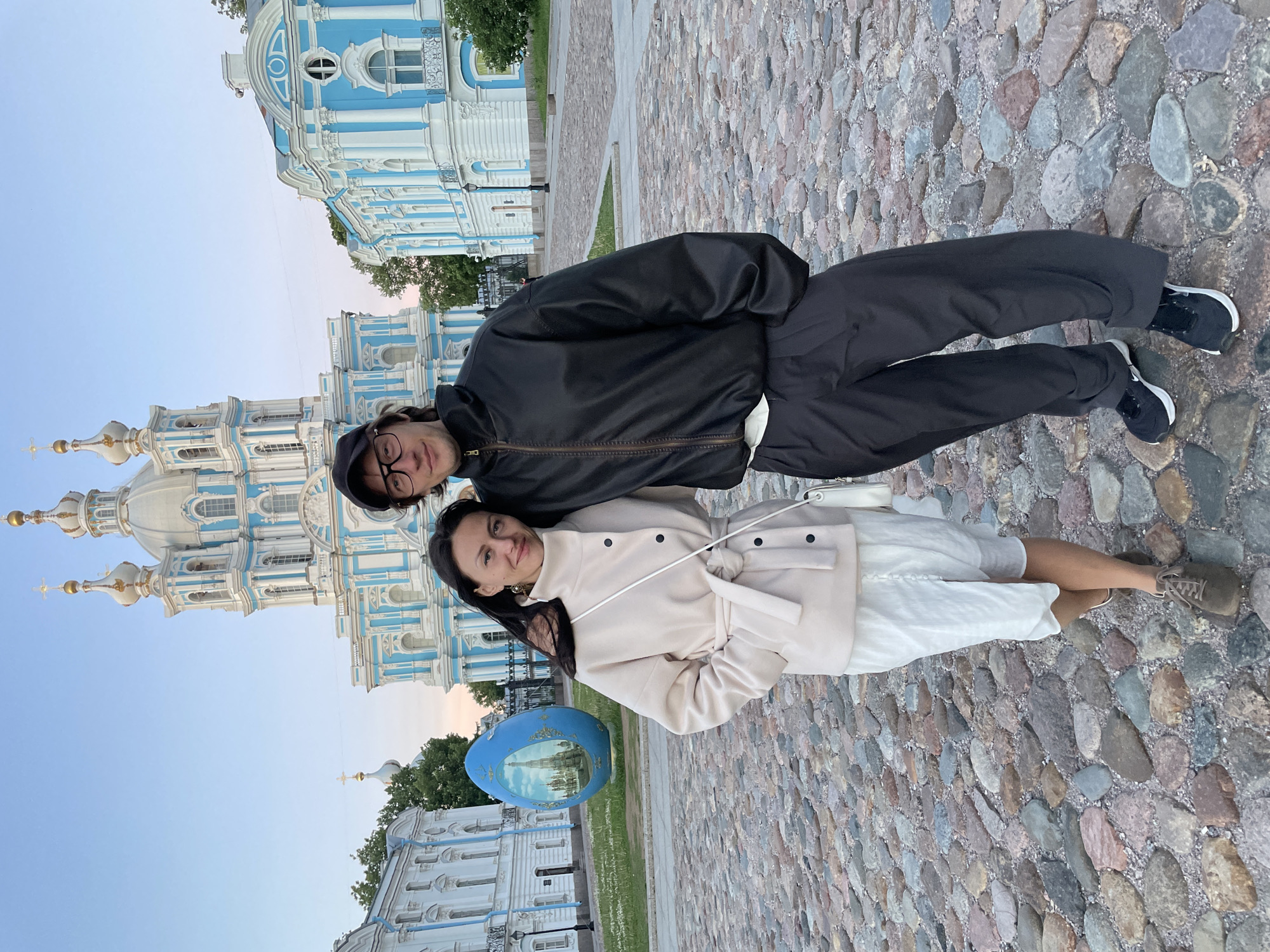 Two individuals, a woman and a man, stand facing the camera on a cobblestone plaza. The woman is on the left, wearing a cream-colored coat and a white skirt. She has dark hair and is looking directly at the camera. The man is on the right, wearing a black jacket and dark pants, with glasses and a black baseball cap. He is also looking at the camera. Behind them is the Smolny Cathedral, a prominent blue and white Baroque cathedral in Saint Petersburg, Russia. To the left of the cathedral, a large, blue, egg-shaped sculpture with an image of a building on it is visible. The sky is clear and pale blue. The overall scene appears to be a daytime photograph taken outdoors in a historical urban setting.